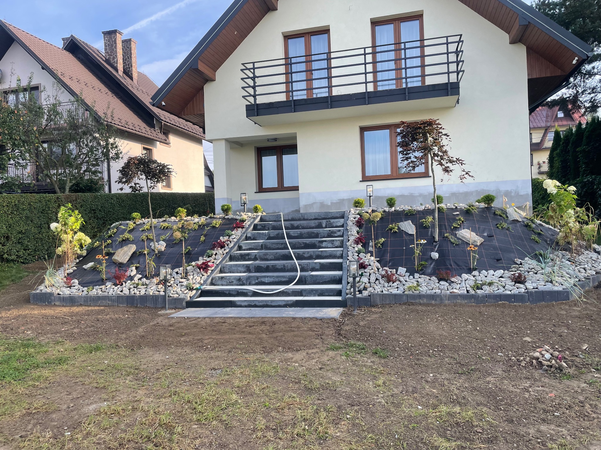 Schody z kostki brukowej prowadzące do domu, obsadzone roślinami i kamieniami ozdobnymi na zboczu, widoczny system nawadniania.