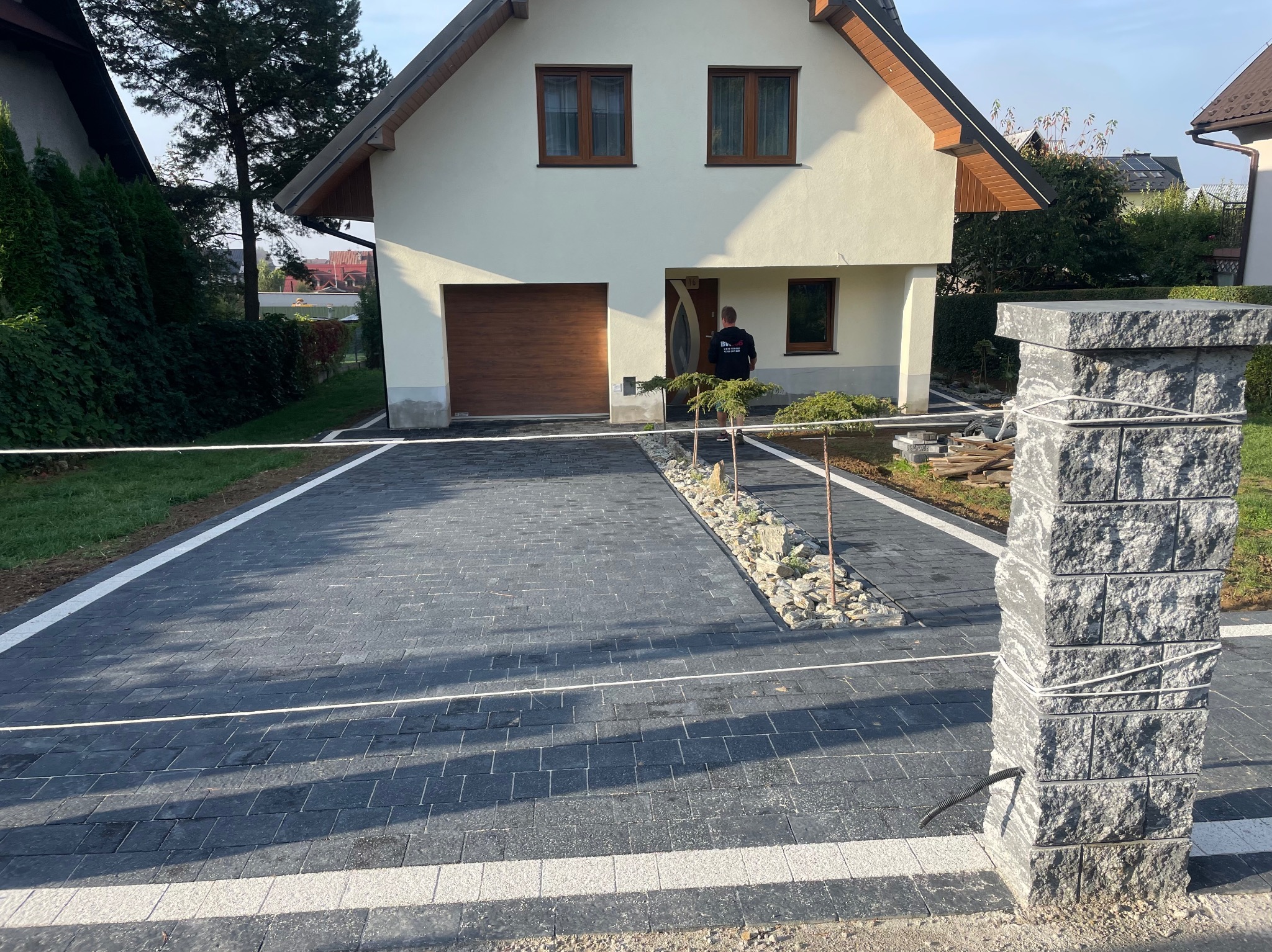 Nowo ułożony, ciemnoszary chodnik z kostki brukowej z białymi obrzeżami prowadzący do domu z brązowym garażem, ozdobiony rabatą z kamieniami i młodymi drzewkami, widoczny fragment słupka...