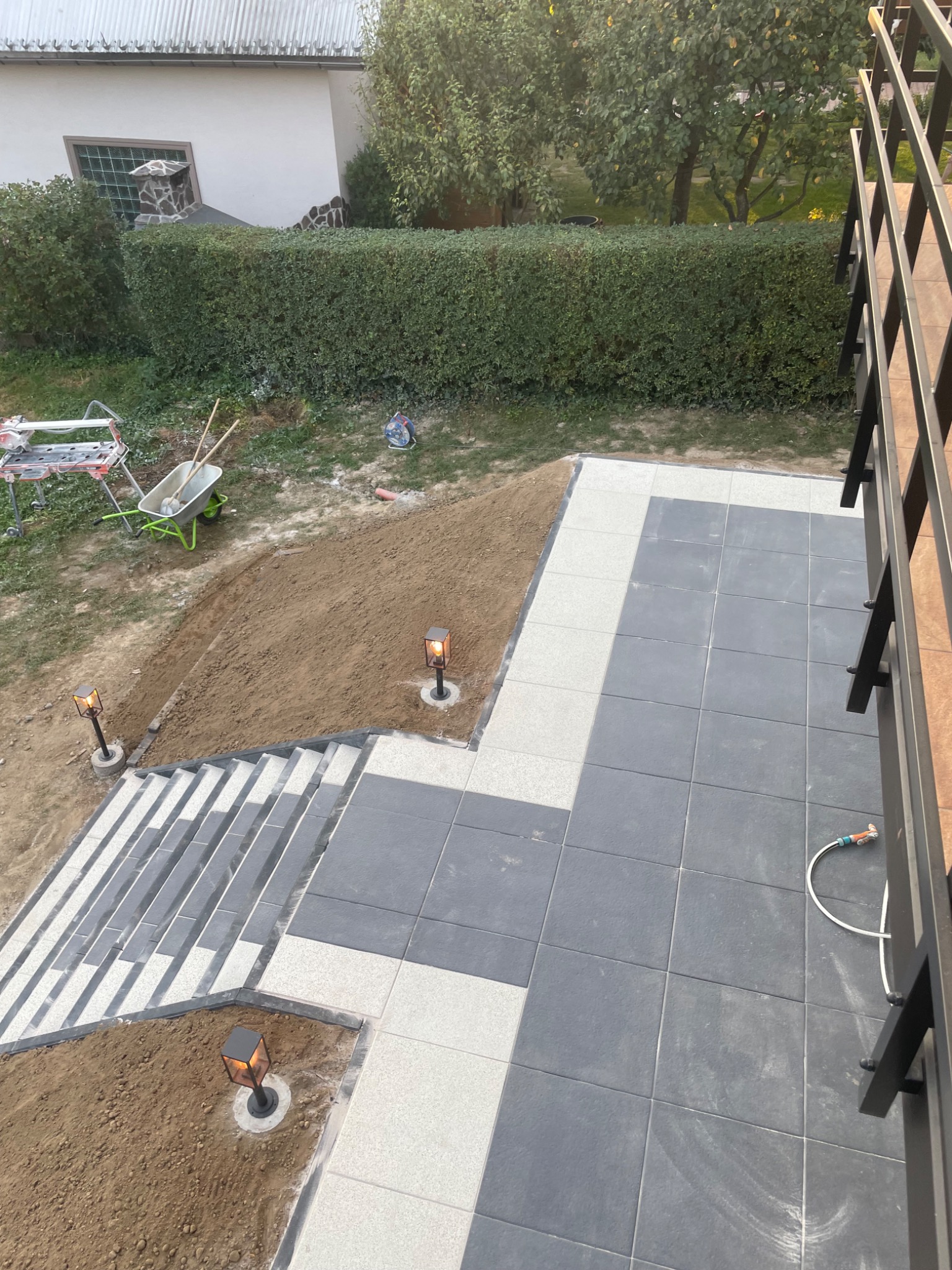 Widok z góry na fragment ogrodu z nową ścieżką z szarych płyt, białą obwódką, schodami z szarych płyt i lampkami ogrodowymi, w tle trawnik, krzewy i narzędzia.