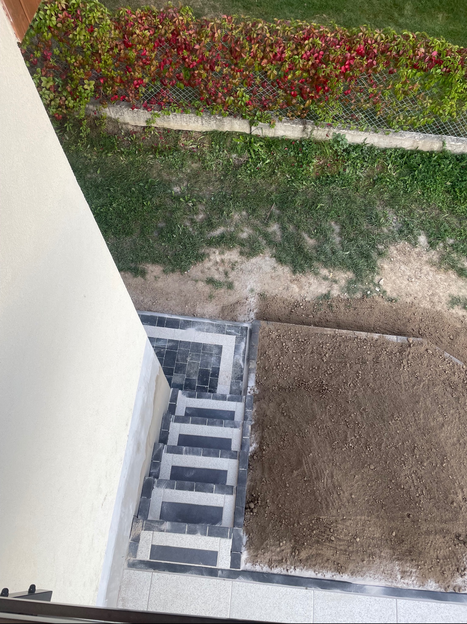 Widok z góry na układanie kostki brukowej przy schodach domu, fragment utwardzonego terenu z przygotowaną podbudową, w tle żywopłot z siatki.