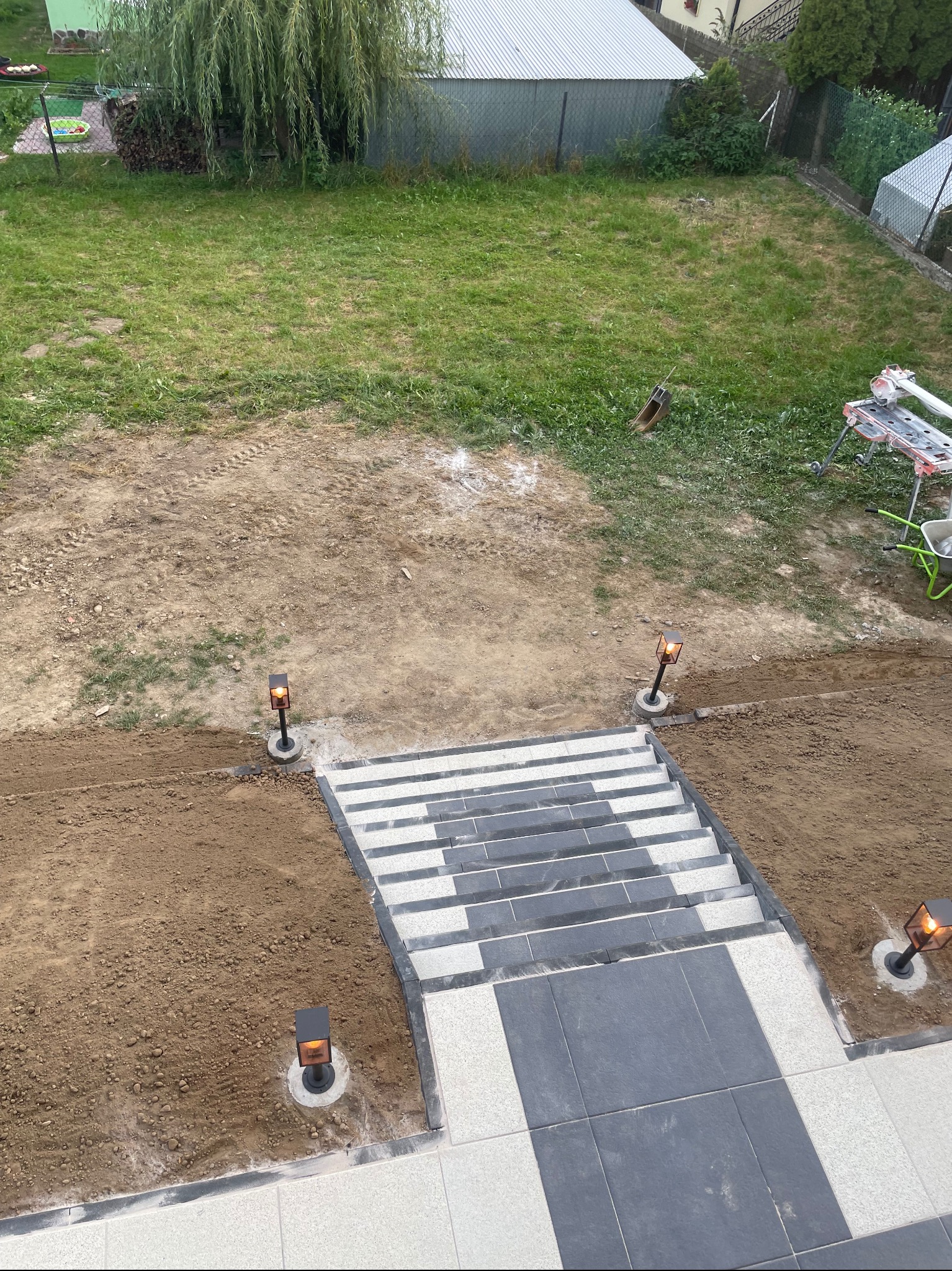 Widok z góry na schody z szarych i białych płyt, otoczone świeżo wyrównaną ziemią i lampami ogrodowymi, z trawnikiem w tle.