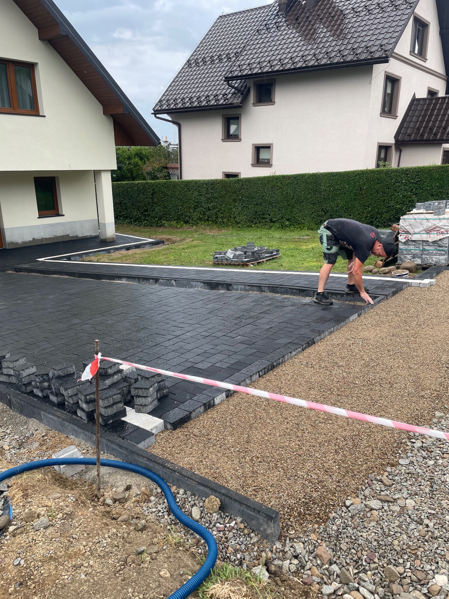 Układanie ciemnej kostki brukowej na posesji, widoczny pracownik w trakcie pracy, przygotowane podłoże z kamyczków, w tle domy i żywopłot.