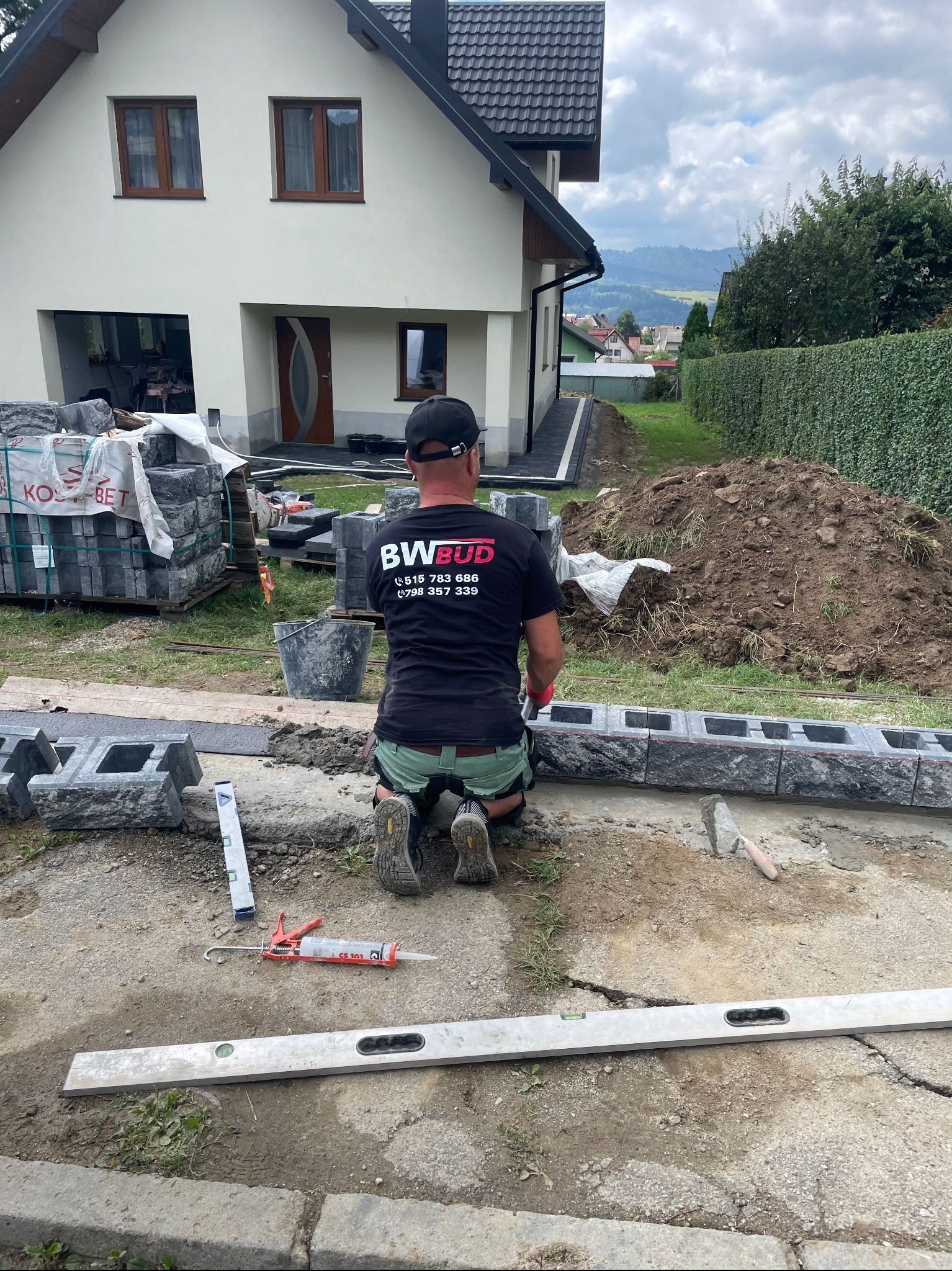 Pracownik w koszulce z logo firmy klęczy układając obrzeża z szarej kostki betonowej przy domu. W tle widoczny stos kostki, narzędzia i pryzma ziemi.