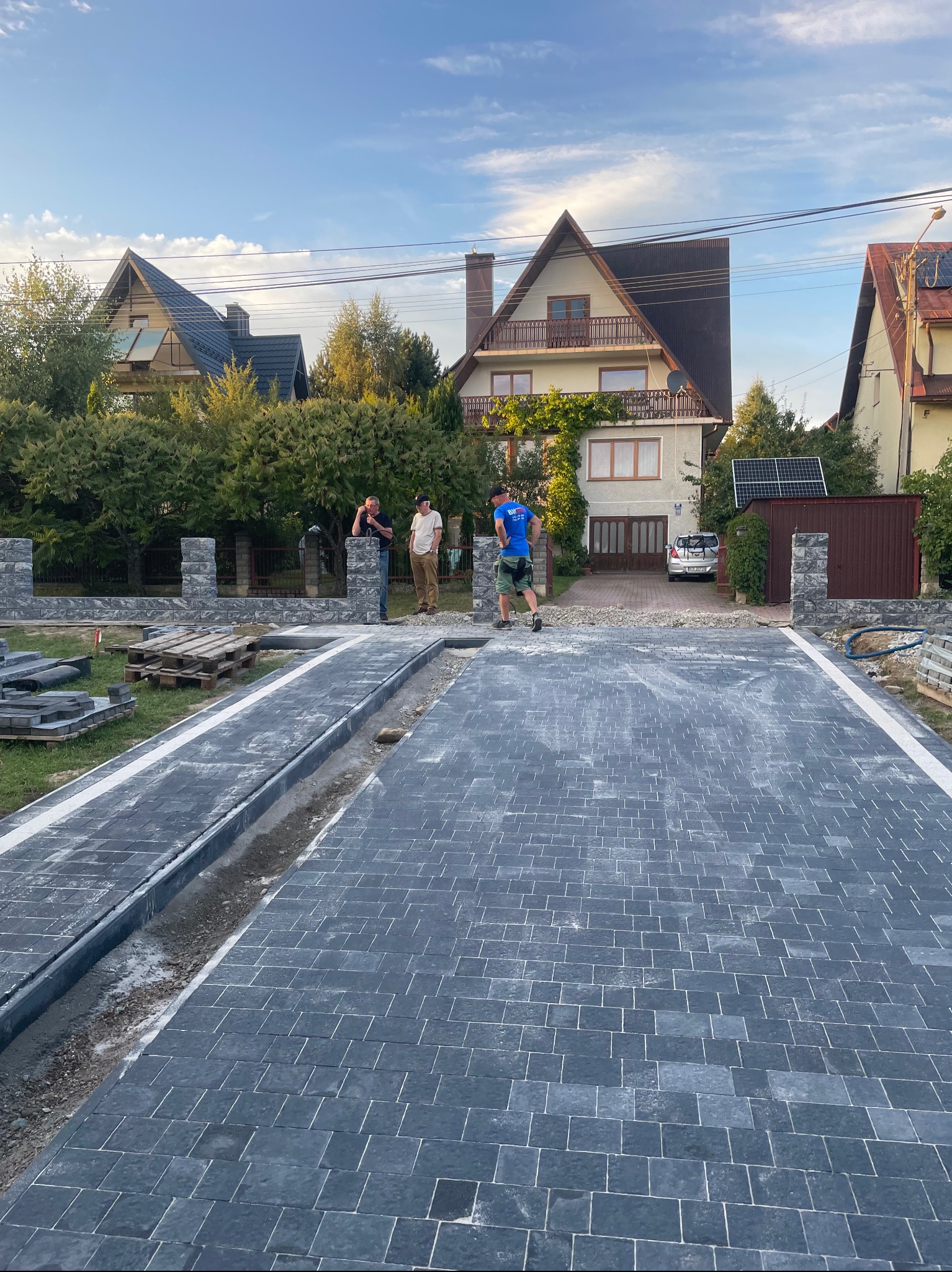 Ciemnoszara kostka brukowa ułożona na podjeździe, widoczny fragment ogrodzenia z kamienia łupanego, w tle domy jednorodzinne i trzech mężczyzn.