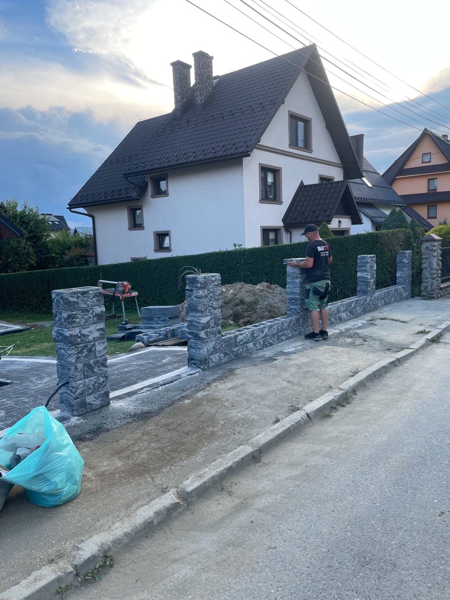 Montaż ogrodzenia z szarej kostki łupanej w Szczawnicy, widoczny pracownik z kostką, w tle dom jednorodzinny i zielony żywopłot.