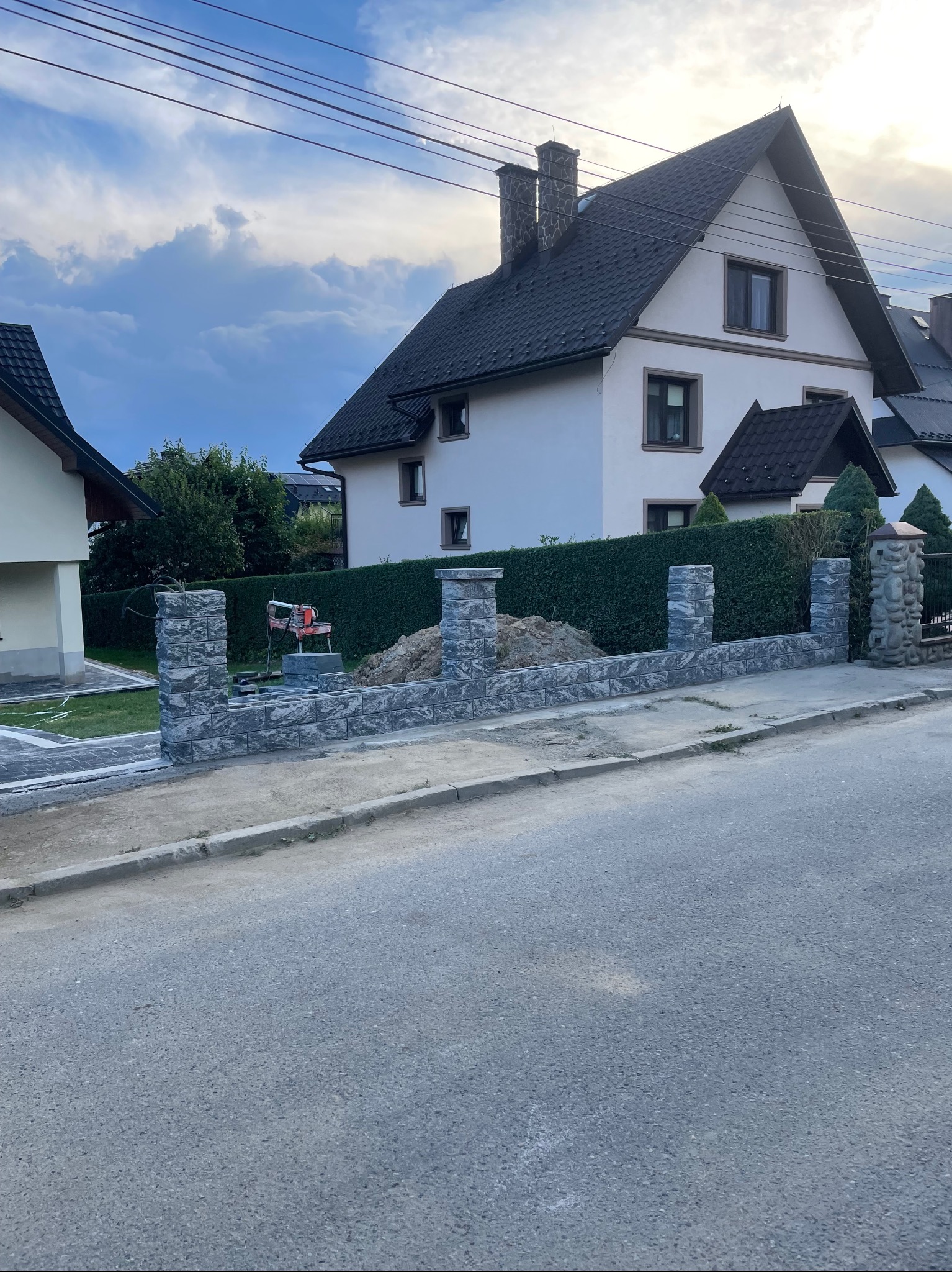 Wznoszenie ogrodzenia z szarej kostki modułowej przed domem z ciemnym dachem, widoczna piła do cięcia kostki i przygotowane podłoże.