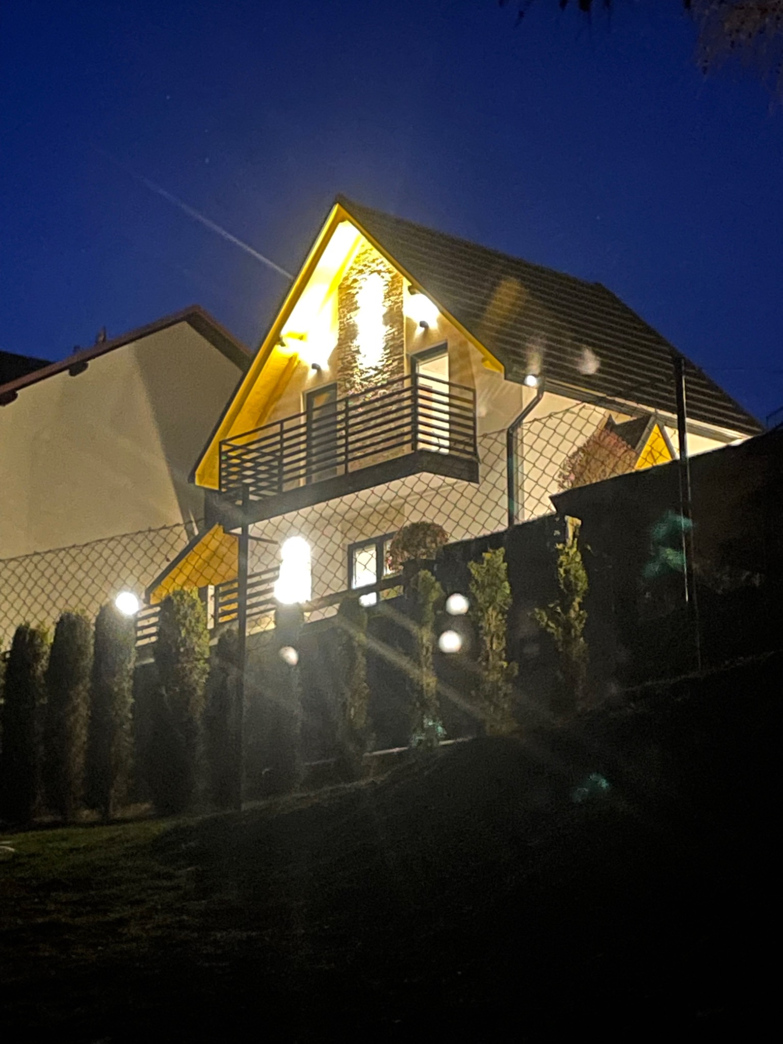 Oświetlony dom jednorodzinny z balkonem, widoczny zza ogrodzenia w nocy, z dodatkowym oświetleniem ogrodowym.