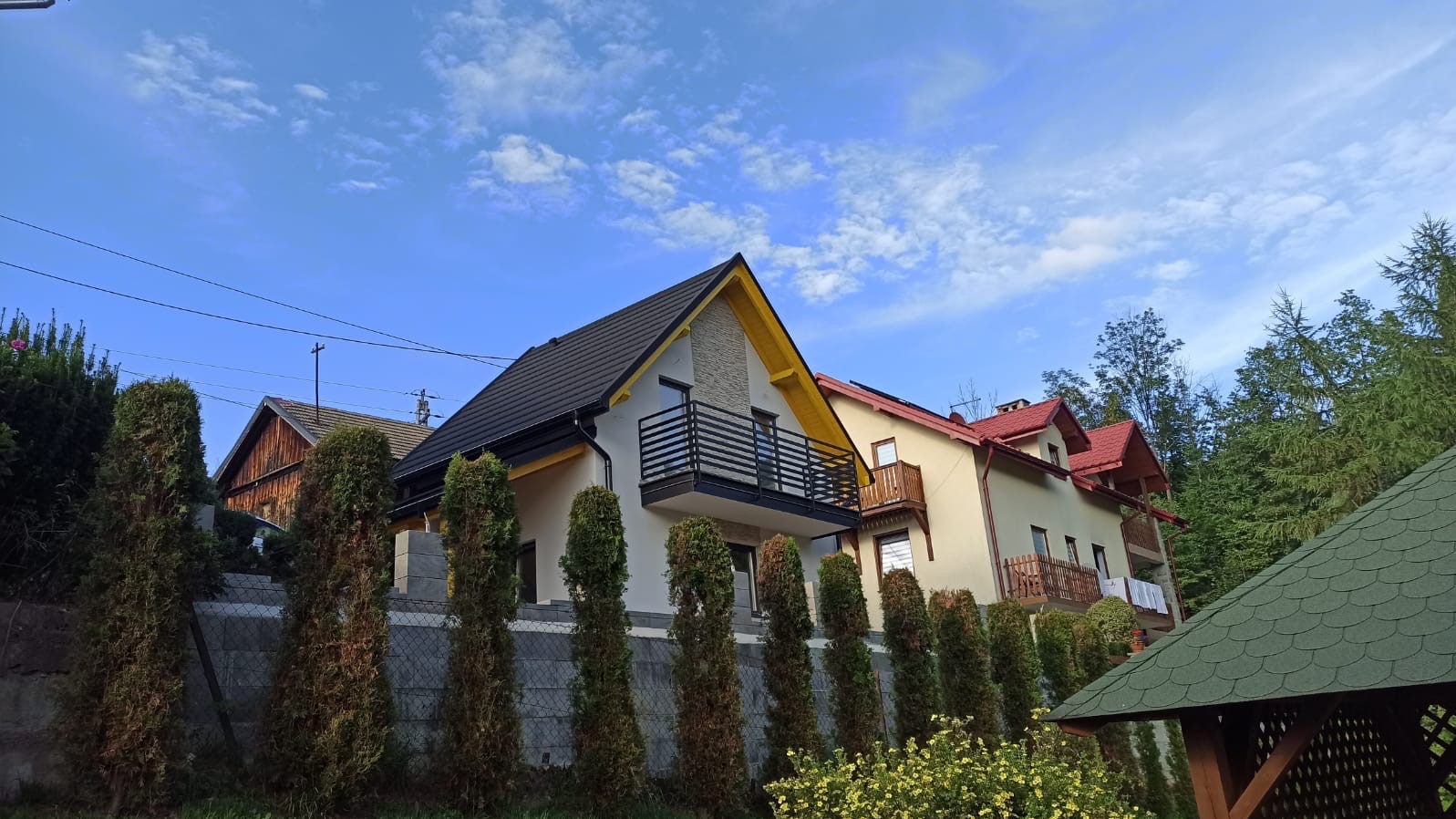 Nowoczesny dom jednorodzinny z czarnym dachem i żółtymi akcentami, balkon z metalową balustradą, tuje wzdłuż ogrodzenia z siatki, błękitne niebo z chmurami.