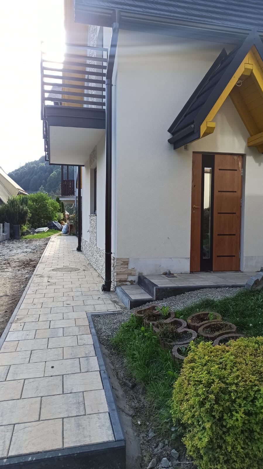 Nowoczesny chodnik z jasnej kostki brukowej prowadzący do domu z balkonem i drewnianymi drzwiami wejściowymi, widok z boku, w tle zieleń i góry.