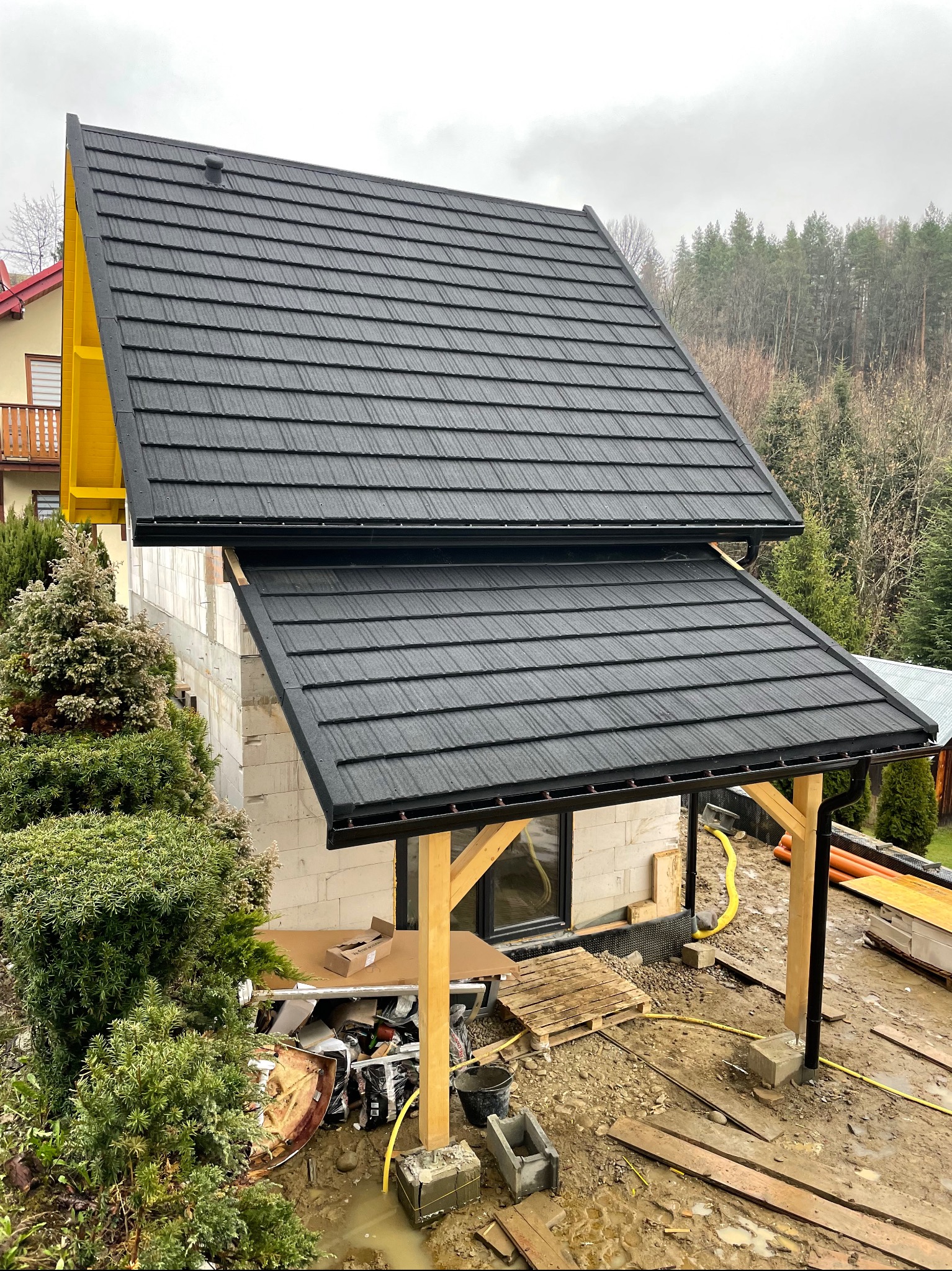 Surowy stan budowy domu z dwuspadowym dachem pokrytym czarną blachodachówką, widoczne drewniane podpory z czarnymi rynnami, gruz i błoto na placu budowy.