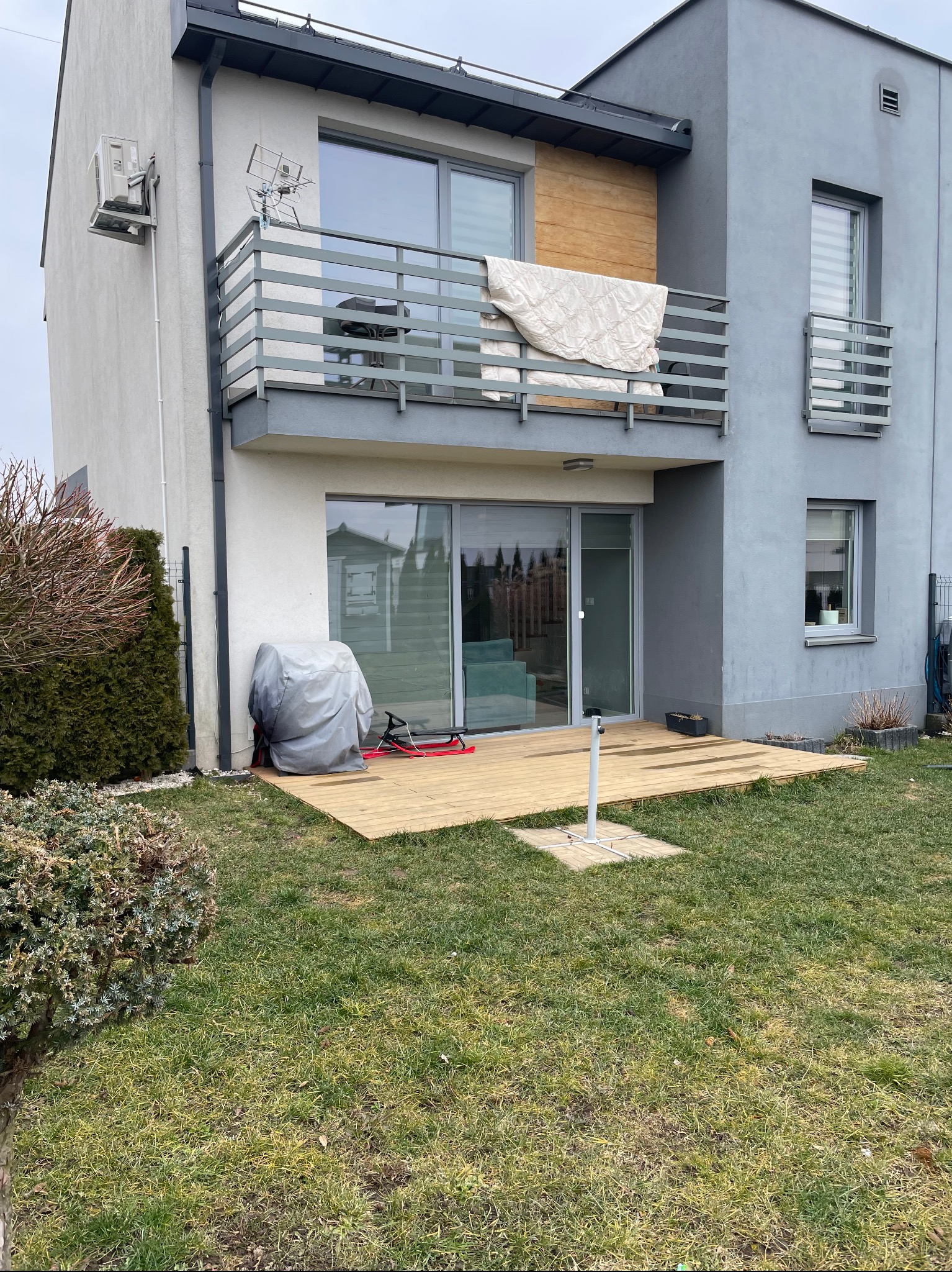 Nowoczesny dom z drewnianym tarasem przylegającym do budynku, widok na trawnik i elementy wyposażenia ogrodu, balkon z meblami i kocem.