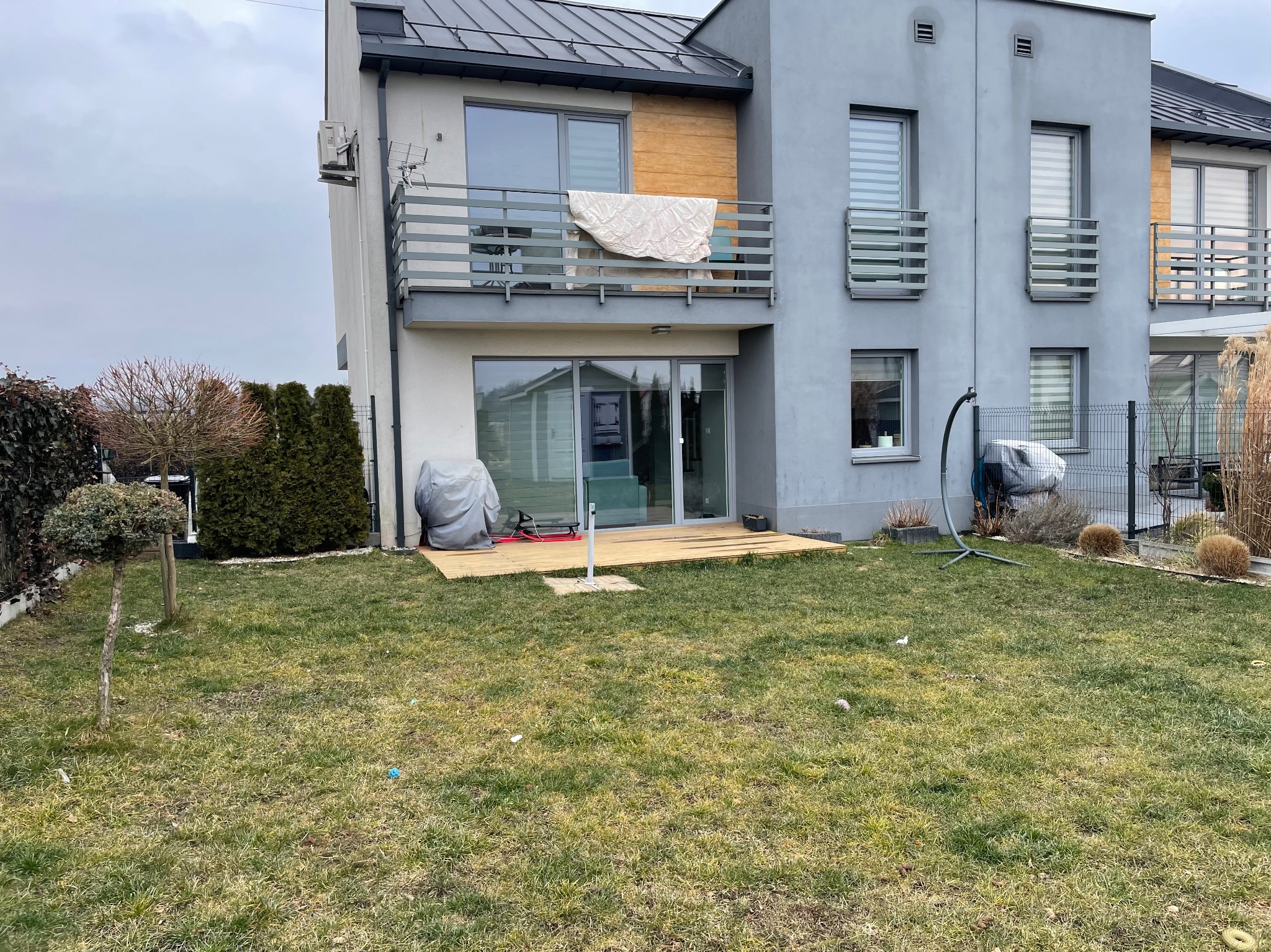 Nowoczesny dom z szarego tynku z drewnianym tarasem, widok z ogrodu. Na balkonie suszy się kołdra, a trawnik jest nieco zaniedbany.