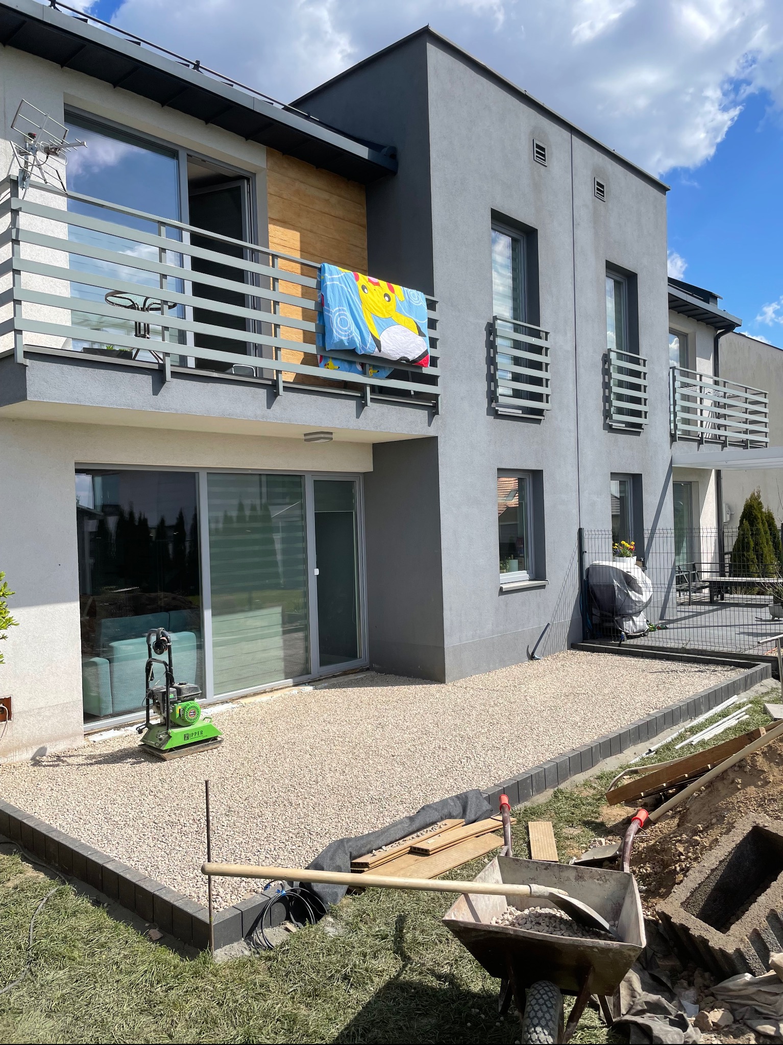 Jasny żwir na posesji przy nowoczesnym domu z balkonami. Widoczna zagęszczarka i taczka z narzędziami. Częściowo ułożona obwódka z kostki brukowej.