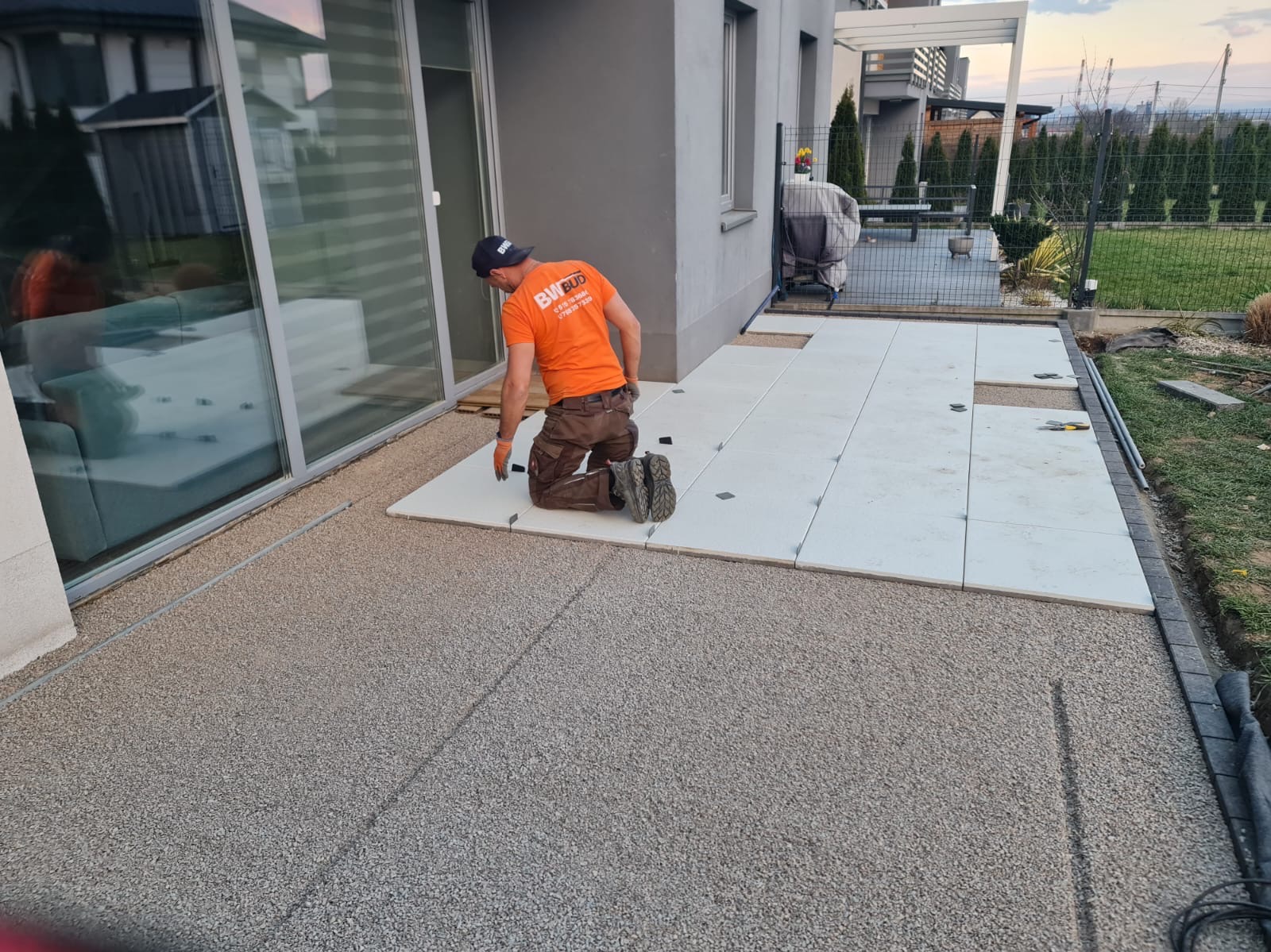 Pracownik w pomarańczowej koszulce klęczy, układając jasne płyty na zewnątrz domu, z widocznym ogrodem i częścią budynku. Płyty ułożone częściowo, widoczny etap prac.