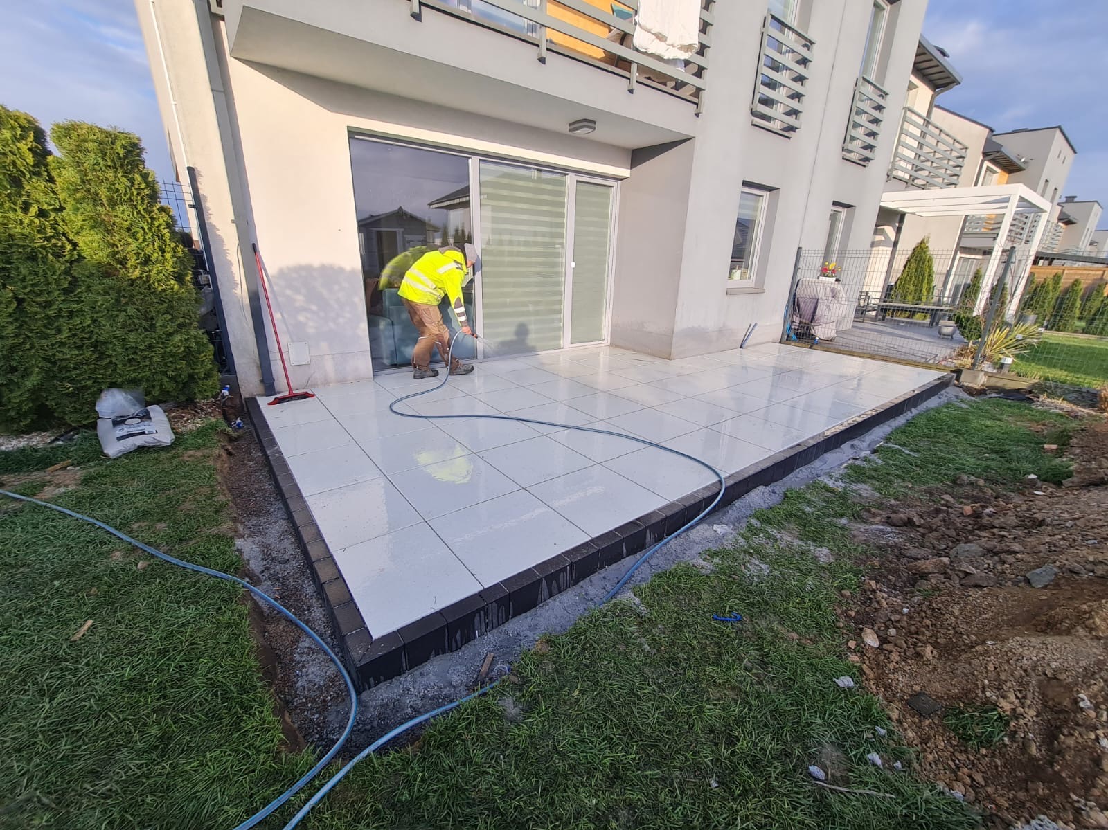 Ułożona jasna kostka na tarasie domu jednorodzinnego, pracownik w żółtej kamizelce czyści powierzchnię. Widoczne obrzeża i przygotowany teren pod trawnik.
