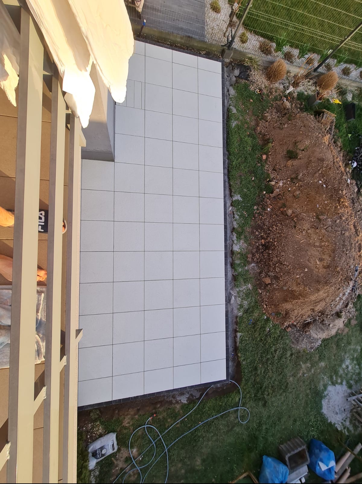 Widok z góry na ułożoną, jasną kostkę brukową na tarasie, z fragmentem budynku i ogrodu. W tle widoczna ziemia i trawa. Kąt ujęcia podkreśla regularność wzoru kostki.