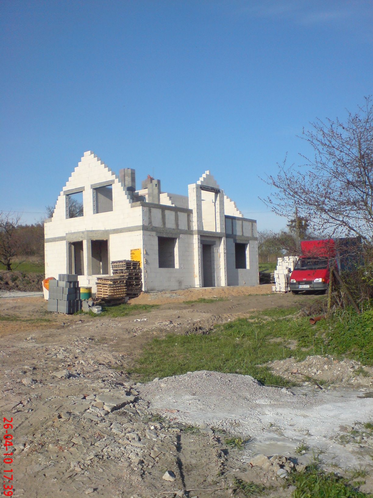 Surowy stan dwukondygnacyjnego domu z białych bloczków, z widocznymi otworami okiennymi i drzwiowymi, na placu budowy z czerwonym samochodem dostawczym w tle.