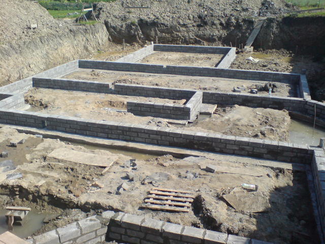 Wykop pod fundamenty z częściowo wzniesionymi ścianami z bloczków betonowych, widoczne drewniane palety na dnie wykopu i ziemia.