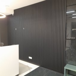 STOLMARK Marek Jakubczyk - Ściana z czarnych, pionowych paneli dekoracyjnych, z widocznym fragmentem białej recepcji i czarną marmurową ścianą po prawej stronie.