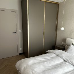 STOLMARK Marek Jakubczyk - Nowoczesna szafa przesuwna w sypialni z łóżkiem i lampką nocną. Szafa w kolorze taupe z złotymi akcentami, dopasowana do drzwi i podłogi w jodełkę.
