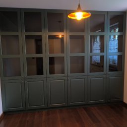 STOLMARK Marek Jakubczyk - Zielona, zabudowana szafa wnękowa z przeszklonymi górnymi drzwiczkami i ciemnymi uchwytami, oświetlona wiszącą lampą, na tle drewnianej podłogi i okna.