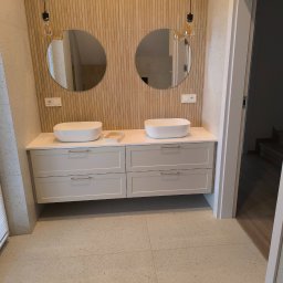 STOLMARK Marek Jakubczyk - Nowoczesna łazienka z podwójną umywalką nablatową, białymi szafkami z szufladami i okrągłymi lustrami pod lampami wiszącymi na tle ściany z drewnopodobnych paneli.