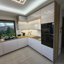 STOLMARK Marek Jakubczyk - Nowoczesna kuchnia w bieli i drewnie z oświetleniem LED, czarnym zlewem i sprzętem AGD. Widok na okno z zielenią i fragment korytarza.