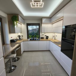 STOLMARK Marek Jakubczyk - Nowoczesna kuchnia z białymi, lakierowanymi frontami i drewnianymi akcentami. Uwagę przykuwa designerska lampa sufitowa i rośliny doniczkowe przy oknie.