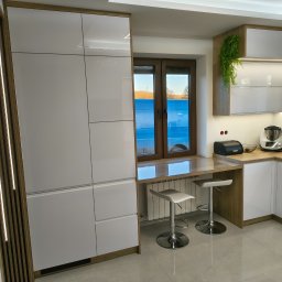 STOLMARK Marek Jakubczyk - Nowoczesna kuchnia z białymi, lakierowanymi frontami, drewnianymi akcentami i oświetleniem LED. Blat roboczy zintegrowany z parapetem, dwa hokery. Widok na zewnątrz.