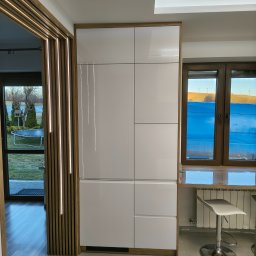 STOLMARK Marek Jakubczyk - Biała, lakierowana szafa w zabudowie z drewnianymi akcentami, w nowoczesnej kuchni. Widoczne okno z blatem i krzesłem barowym. Dekoracyjne lamele przy wejściu.