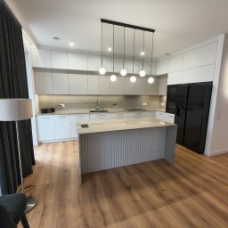 STOLMARK Marek Jakubczyk - Nowoczesna kuchnia z wyspą: białe szafki, marmurowy blat, czarna lodówka side-by-side i designerskie lampy wiszące. Parkiet dodaje ciepła.