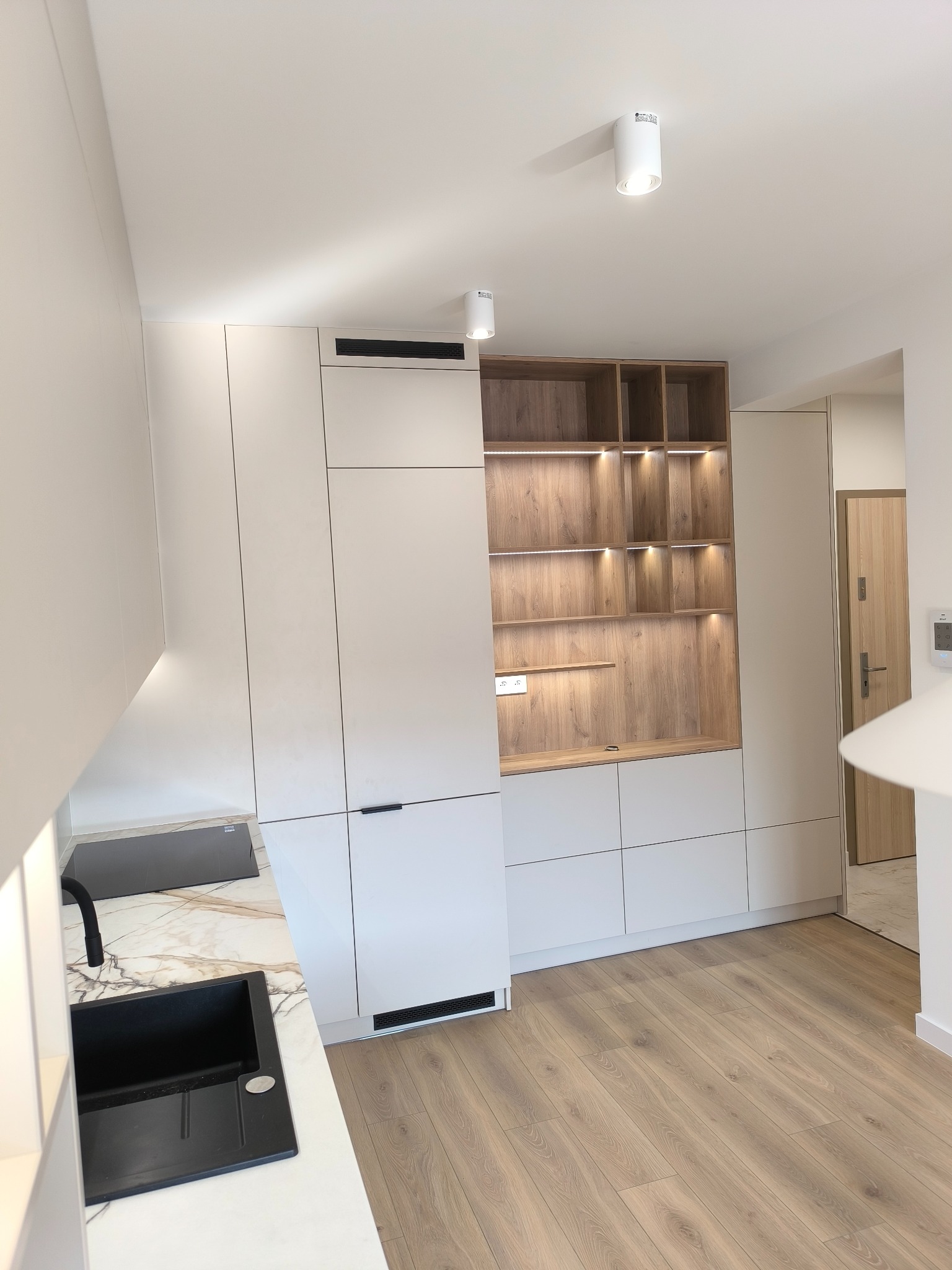 Jasna kuchnia z białymi frontami i drewnianymi akcentami. Otwarta szafka z podświetleniem, czarny zlew na blacie imitującym marmur, parkiet. Minimalistyczny design.