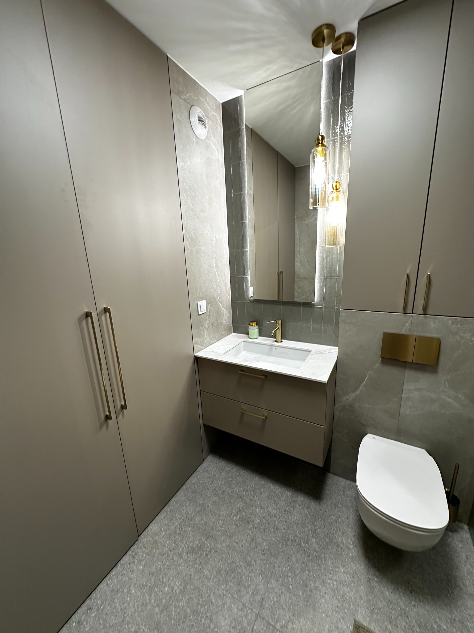 Nowoczesna łazienka z beżowymi szafkami, złotymi uchwytami i marmurowymi płytkami. Wisząca toaleta i lustro z lampami dodają elegancji.