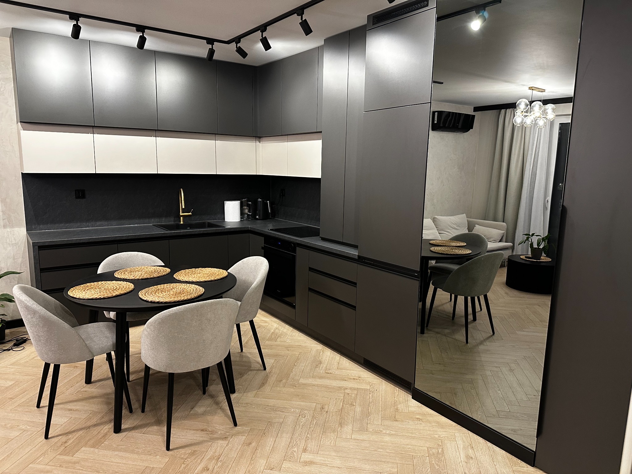 Nowoczesna kuchnia z szarymi szafkami, białymi akcentami i czarnym blatem. Okrągły stół z szarymi krzesłami. W lustrzanej ścianie odbicie salonu.