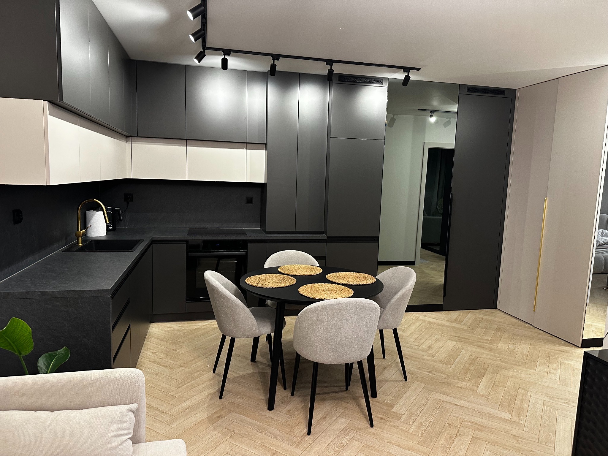 Nowoczesna kuchnia w odcieniach szarości i bieli z czarnym blatem, okrągłym stołem i czterema krzesłami. Widoczne oświetlenie punktowe i parkiet ułożony w jodełkę.