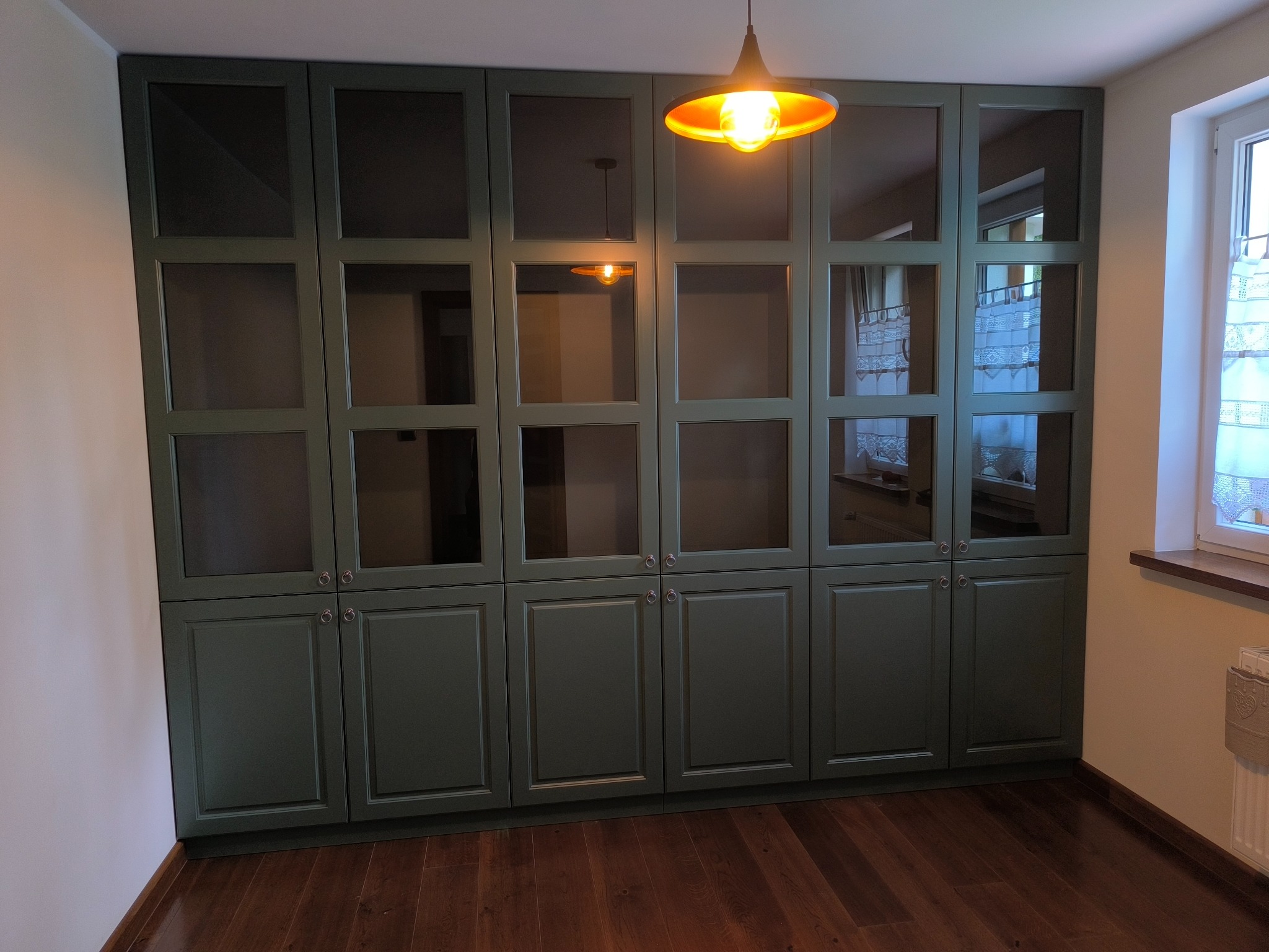 Zielona, zabudowana szafa wnękowa z przeszklonymi górnymi drzwiczkami i ciemnymi uchwytami, oświetlona wiszącą lampą, na tle drewnianej podłogi i okna.