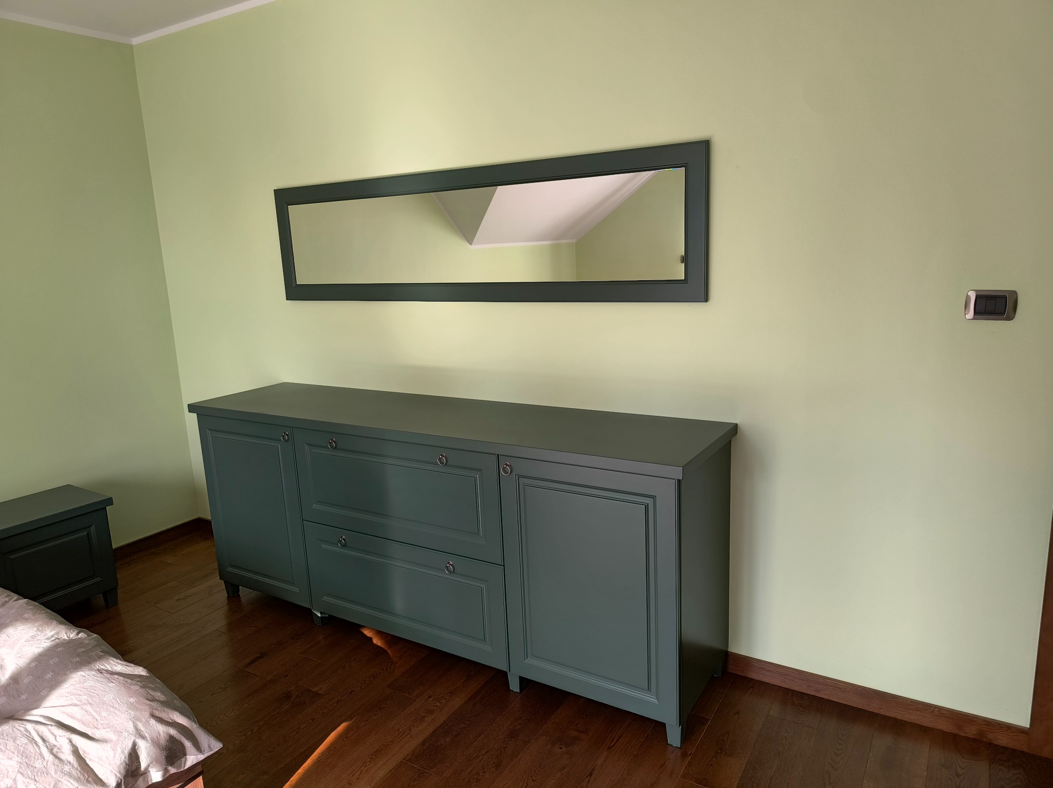 Szara komoda z szufladami i szafkami, z długim lustrem w ramie na ścianie, w pokoju o pistacjowych ścianach i drewnianej podłodze.