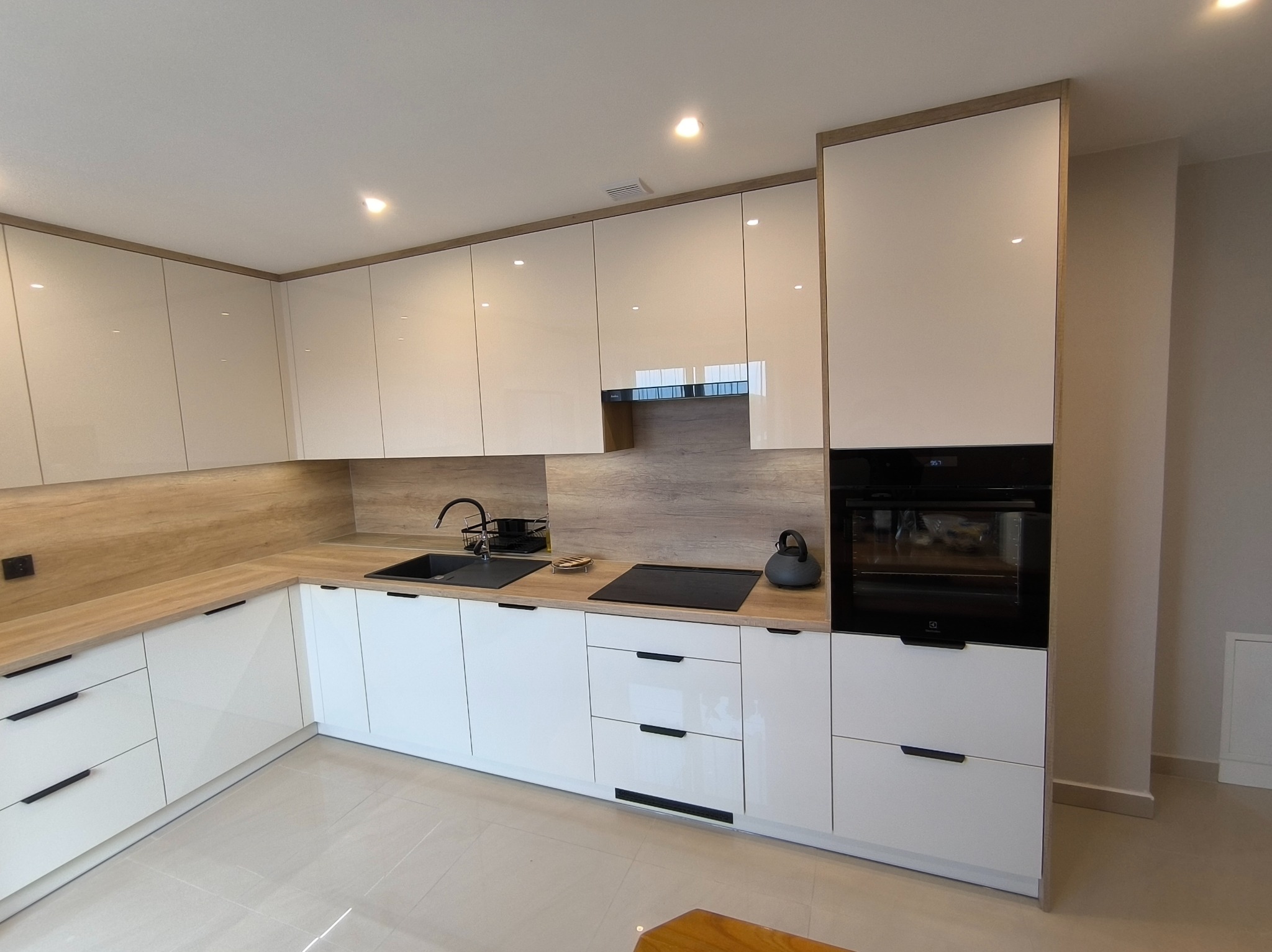 Nowoczesna kuchnia z białymi, lakierowanymi frontami i drewnianym blatem. Minimalistyczne uchwyty, czarny zlew i płyta indukcyjna. Oświetlenie punktowe.