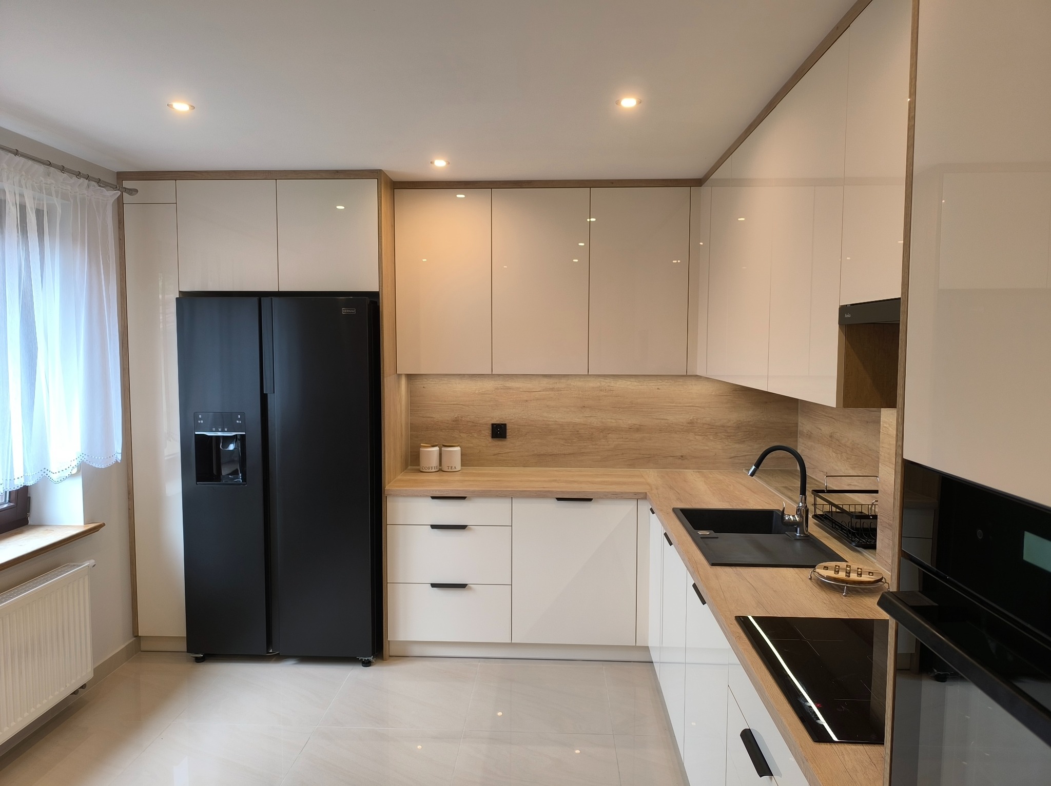 Nowoczesna kuchnia: białe, lakierowane fronty szafek, drewniany blat, czarny zlew i lodówka side-by-side. Minimalistyczny design z subtelnym oświetleniem punktowym.