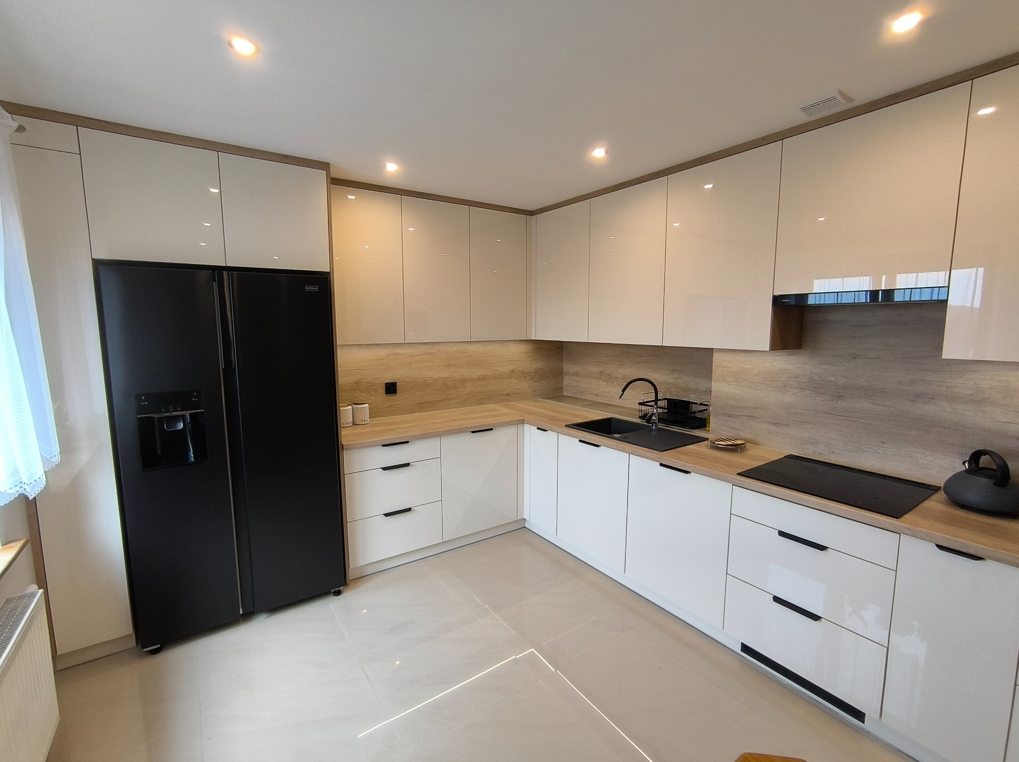 Nowoczesna kuchnia z białymi, lakierowanymi frontami, drewnianym blatem i czarną lodówką side-by-side. Oświetlenie punktowe podkreśla minimalistyczny design.