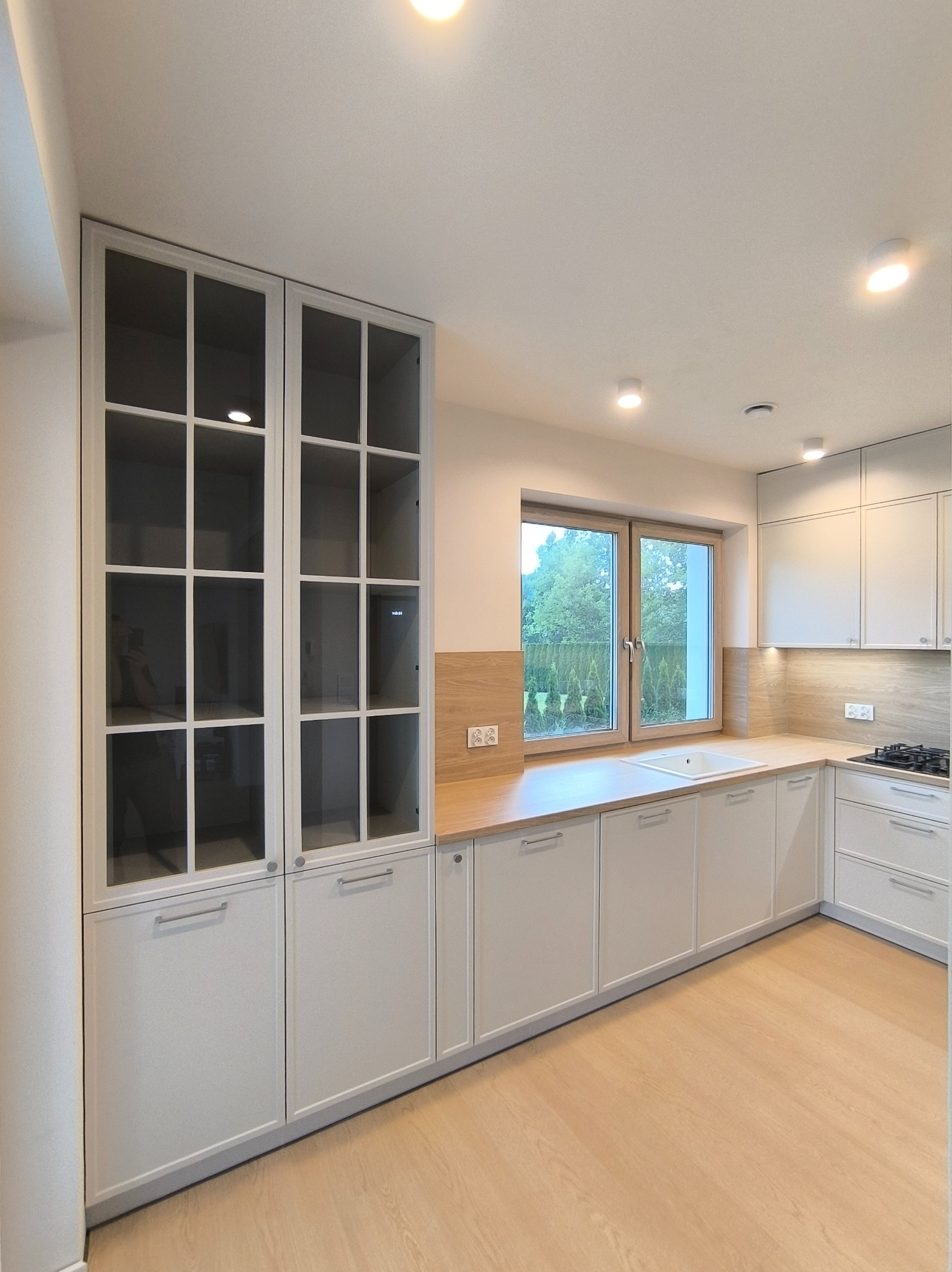 Jasna kuchnia z przeszkloną witryną i drewnianym blatem, oświetlona punktowo, z widokiem na zieleń za oknem. Minimalistyczny design.