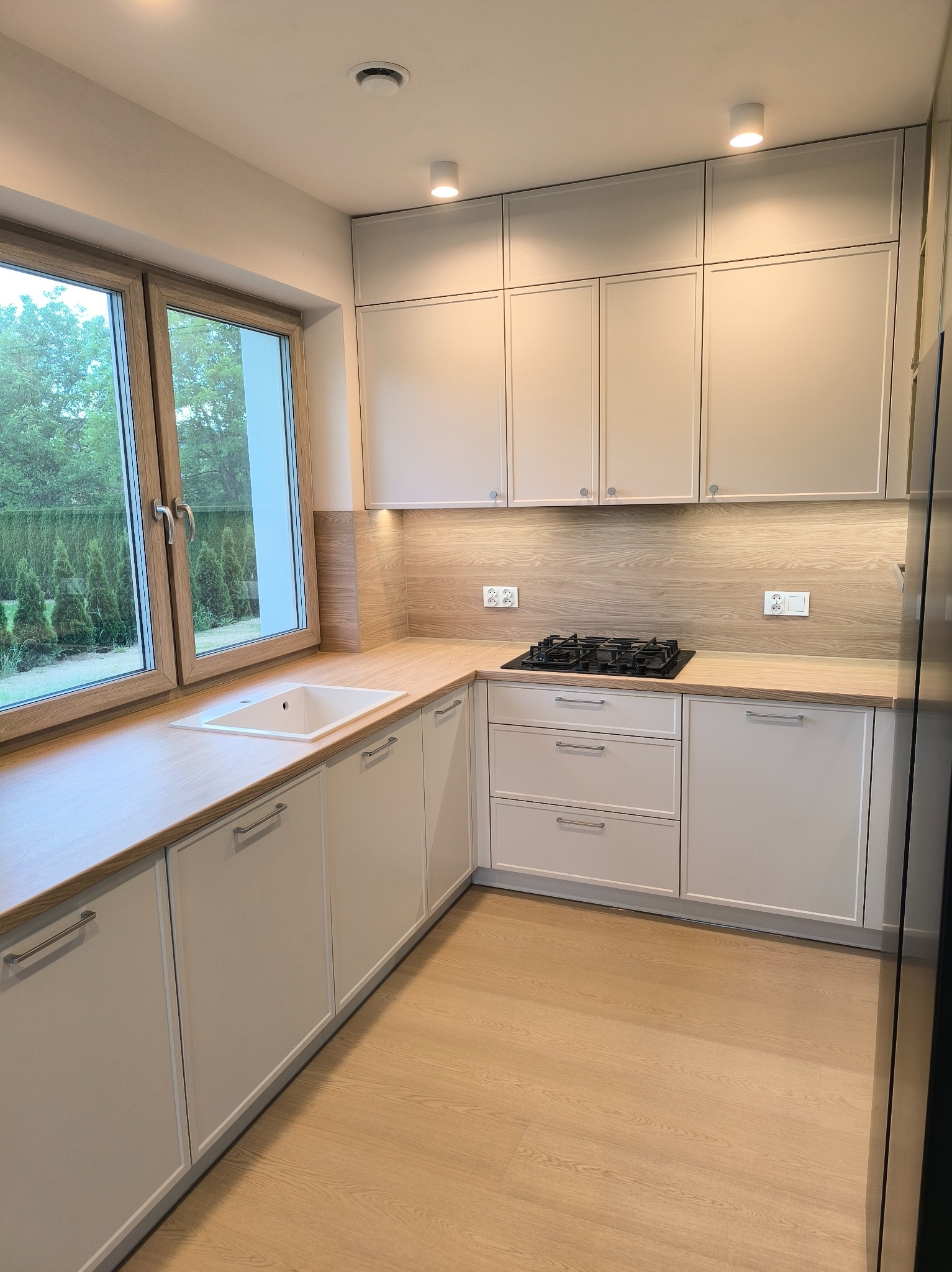 Jasna kuchnia na wymiar z drewnianym blatem i białymi frontami. Widoczny zlew pod oknem i płyta gazowa. Minimalistyczny design, nowoczesne oświetlenie.