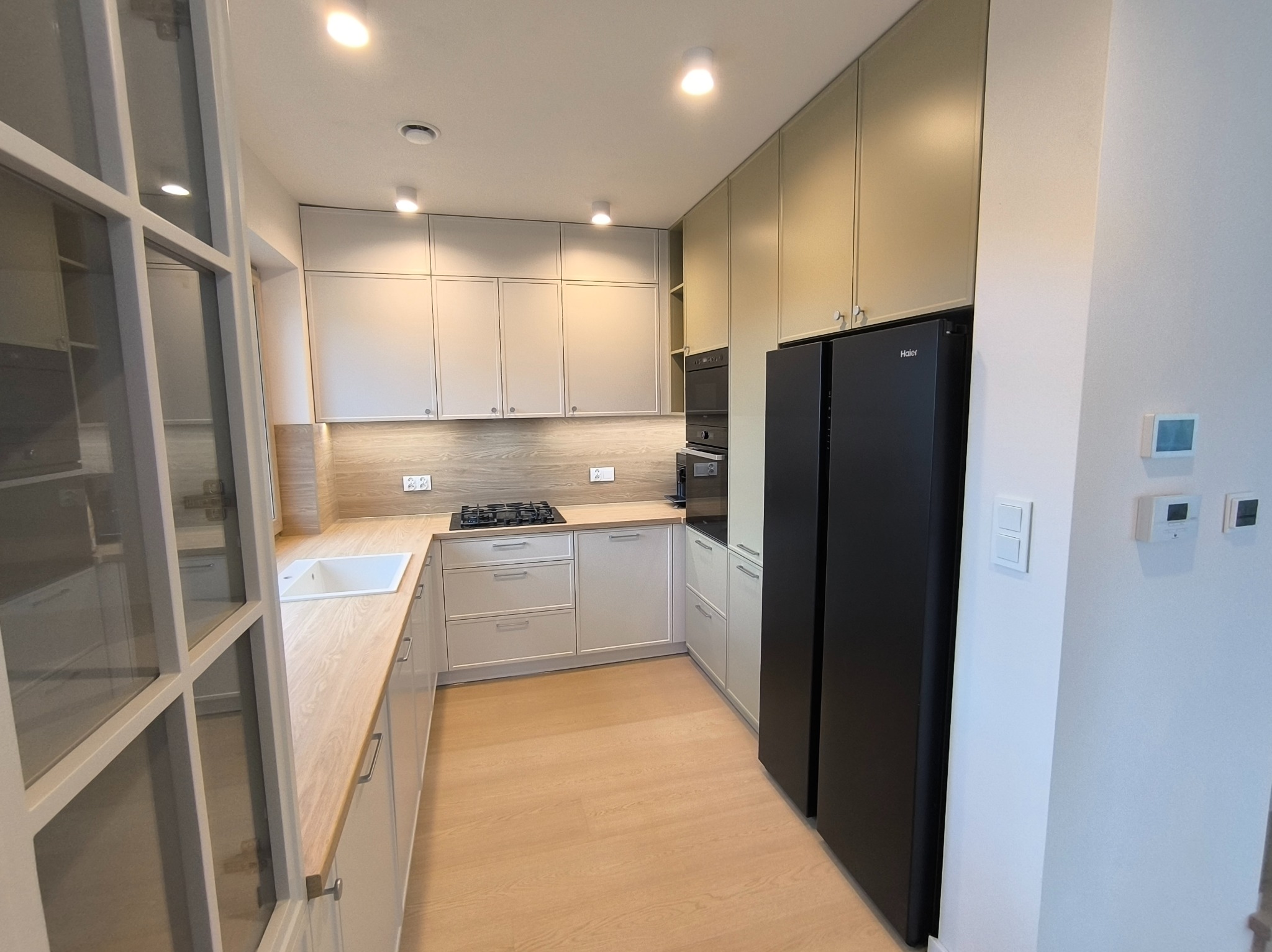 Jasna kuchnia z drewnianym blatem i czarną lodówką. Widoczne białe fronty szafek, płyta grzewcza i okno z podziałami. Nowoczesne oświetlenie punktowe.