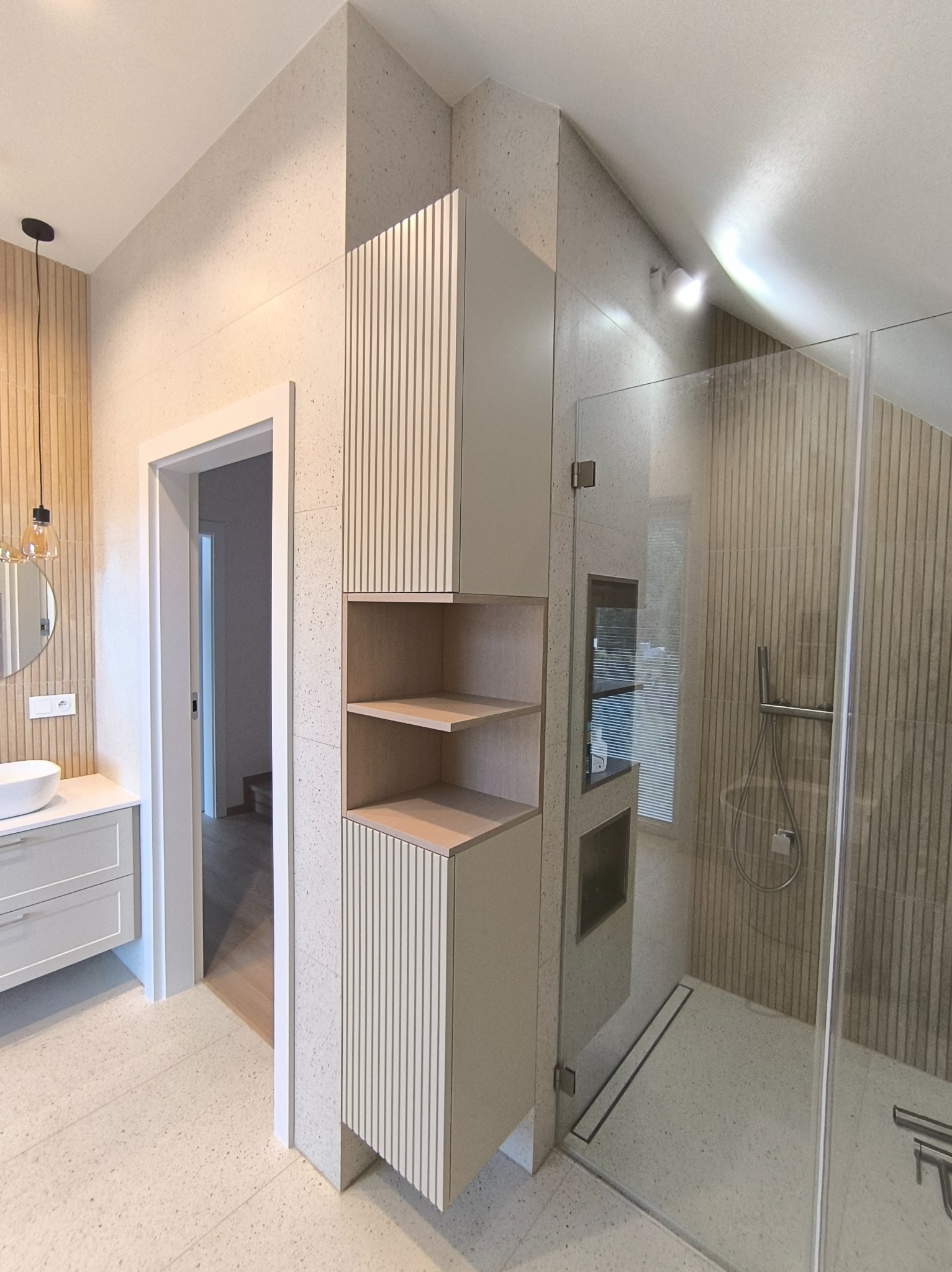 Nowoczesna łazienka z kabiną prysznicową typu walk-in, szafką z półkami i umywalką nablatową. Uwagę zwraca wykończenie ścian i podłogi w jasnym lastryko.