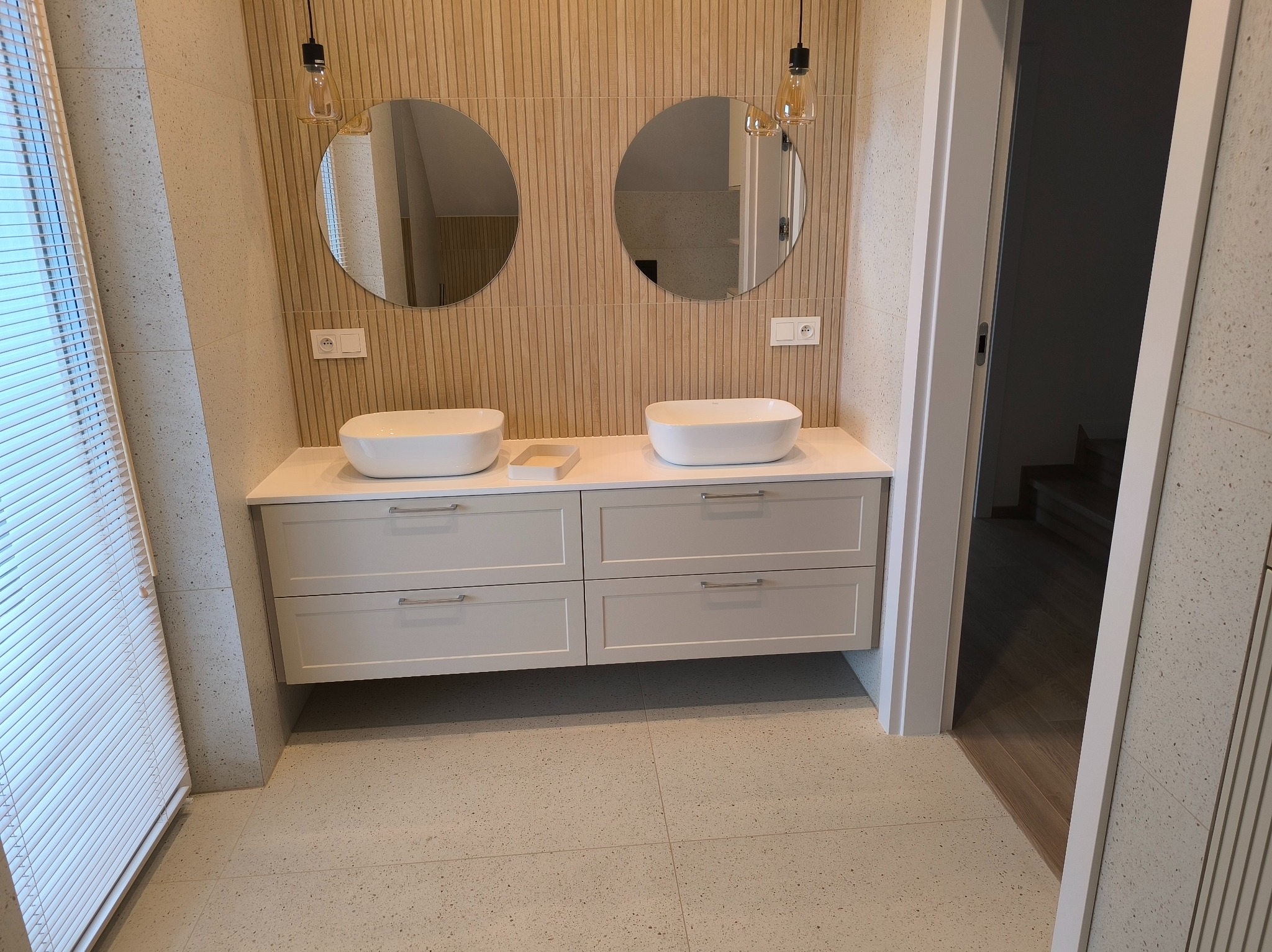 Nowoczesna łazienka z podwójną umywalką nablatową, białymi szafkami z szufladami i okrągłymi lustrami pod lampami wiszącymi na tle ściany z drewnopodobnych paneli.