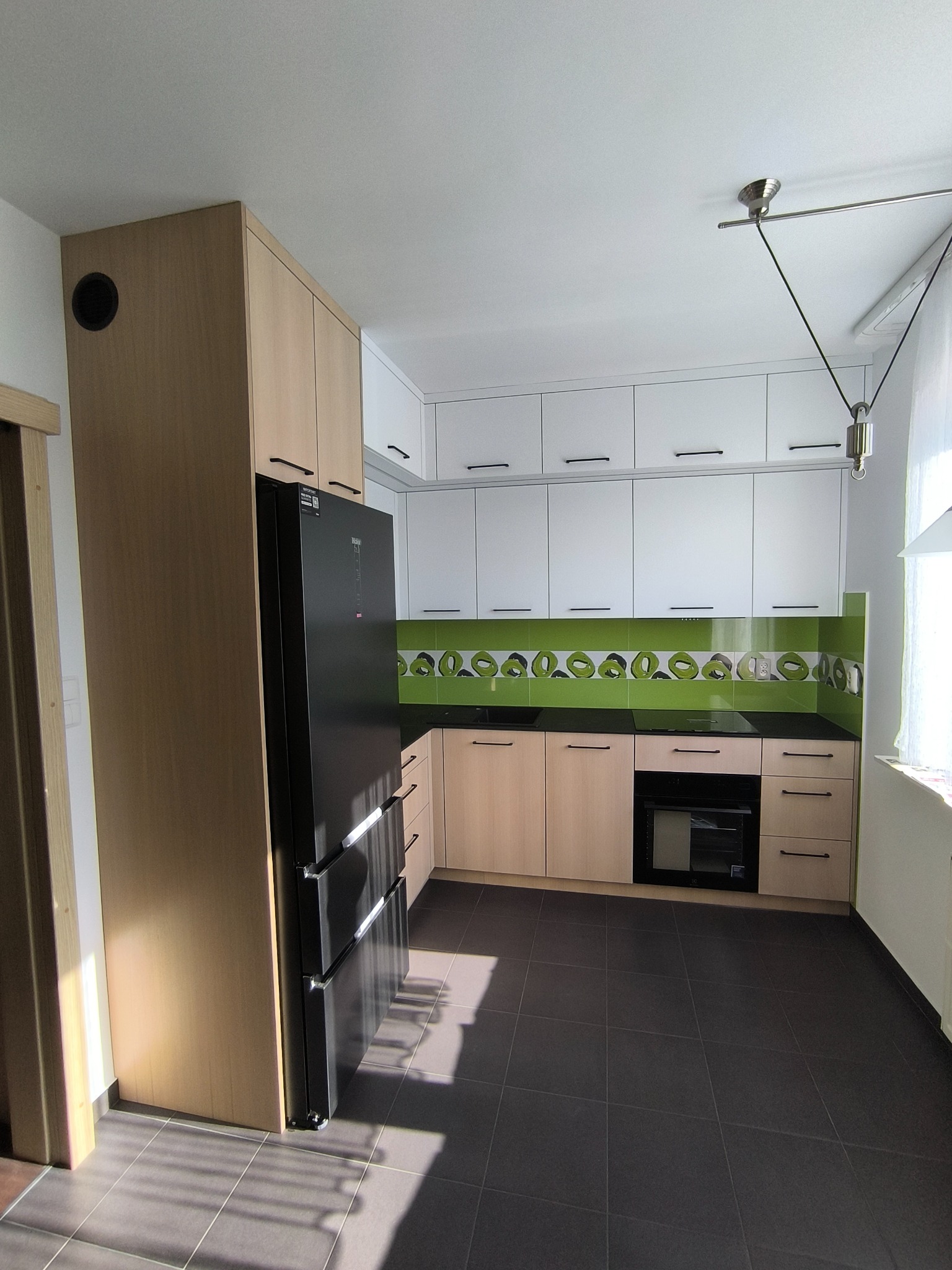 Nowoczesna kuchnia na wymiar: jasne szafki z czarnymi uchwytami, zielony panel nad blatem, czarna lodówka i grafitowa podłoga. Minimalistyczny design z oświetleniem.