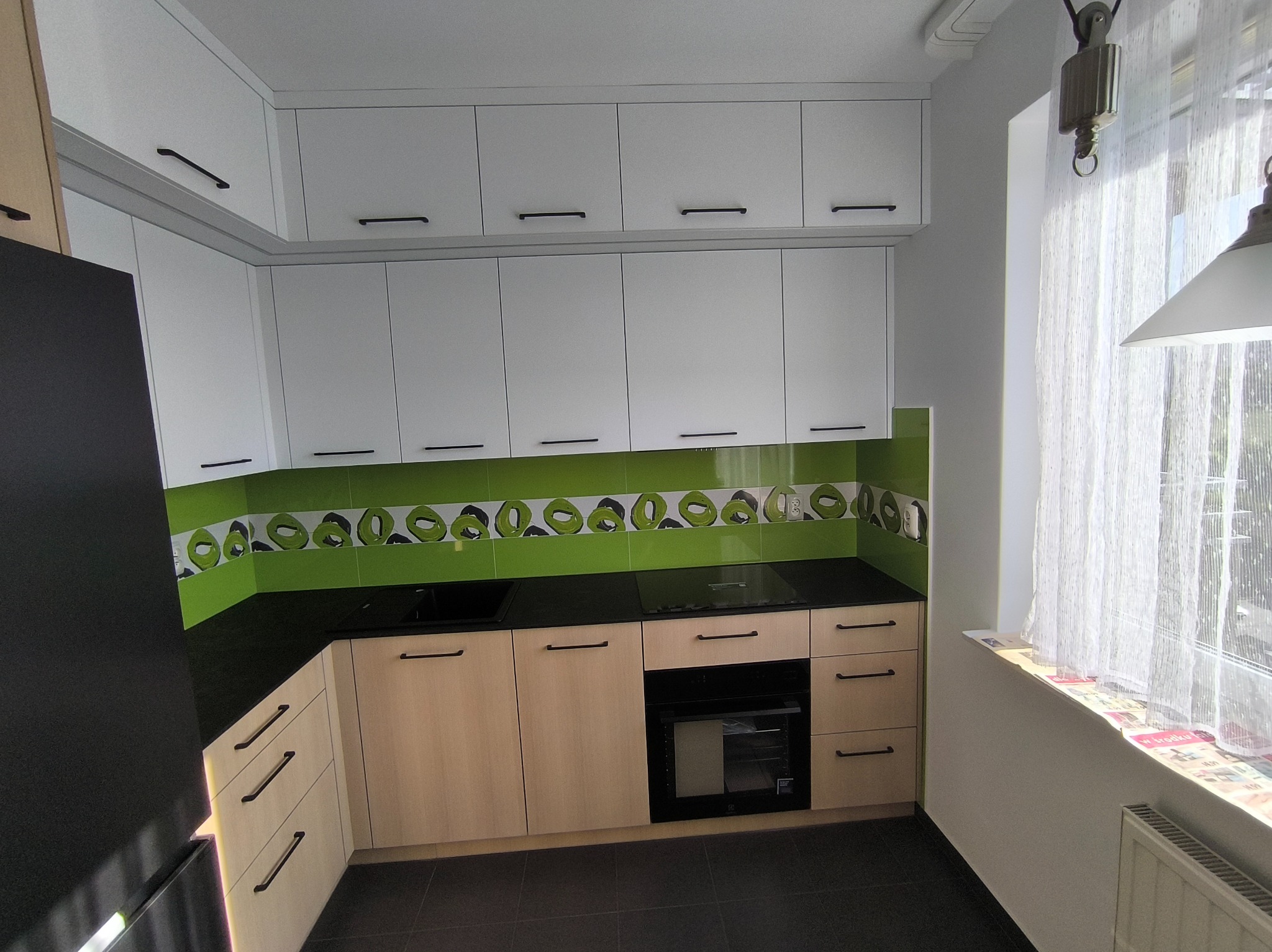 Nowoczesna kuchnia z białymi szafkami górnymi i drewnopodobnymi dolnymi, czarnym blatem oraz zielonymi płytkami z wzorem. Widoczne okno z firanką i czarne uchwyty.