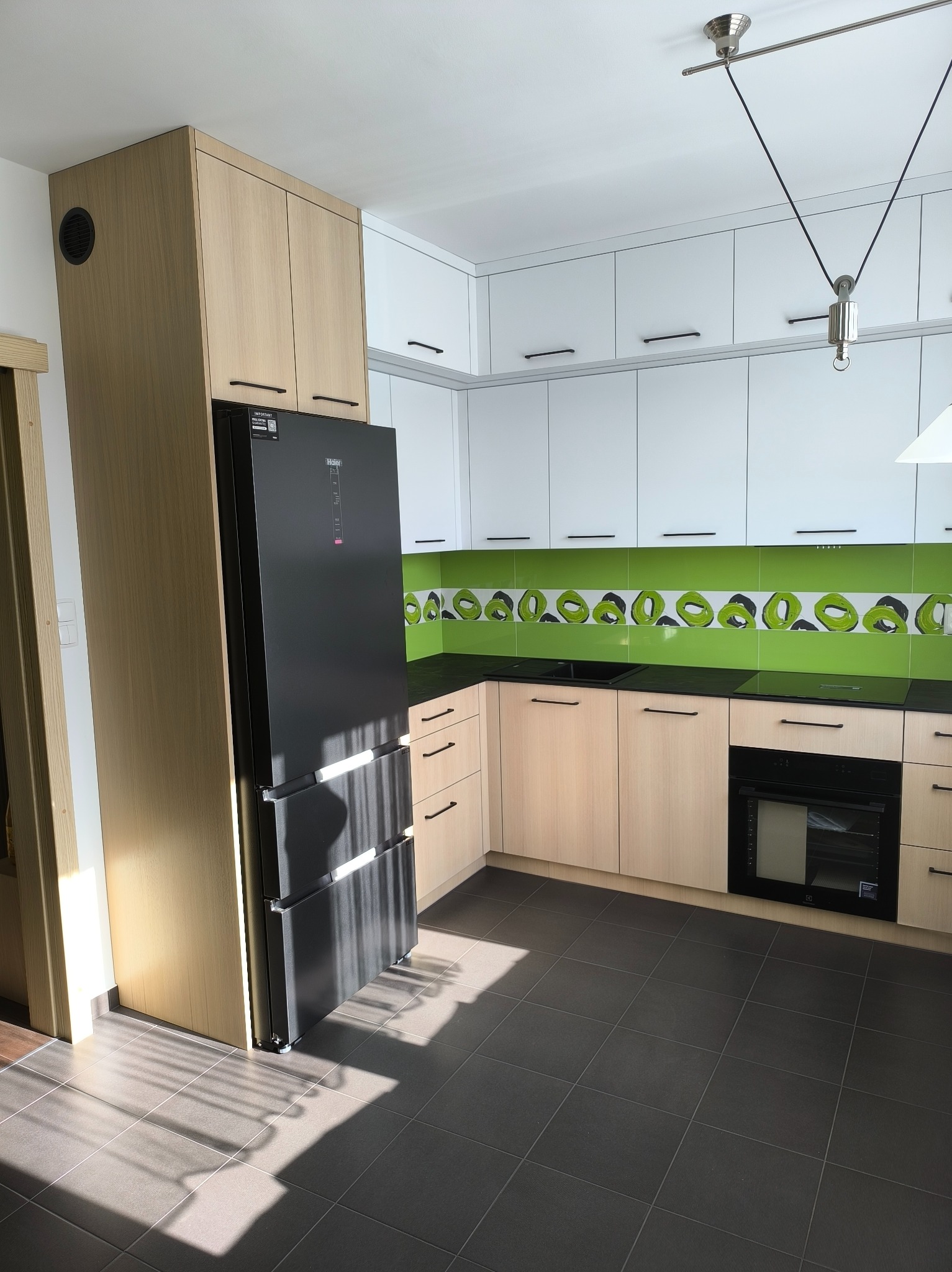 Jasna kuchnia na wymiar: beżowe szafki dolne i wysokie, białe górne, czarny blat i sprzęt AGD, zielony pas płytek. Nowoczesne oświetlenie sufitowe.