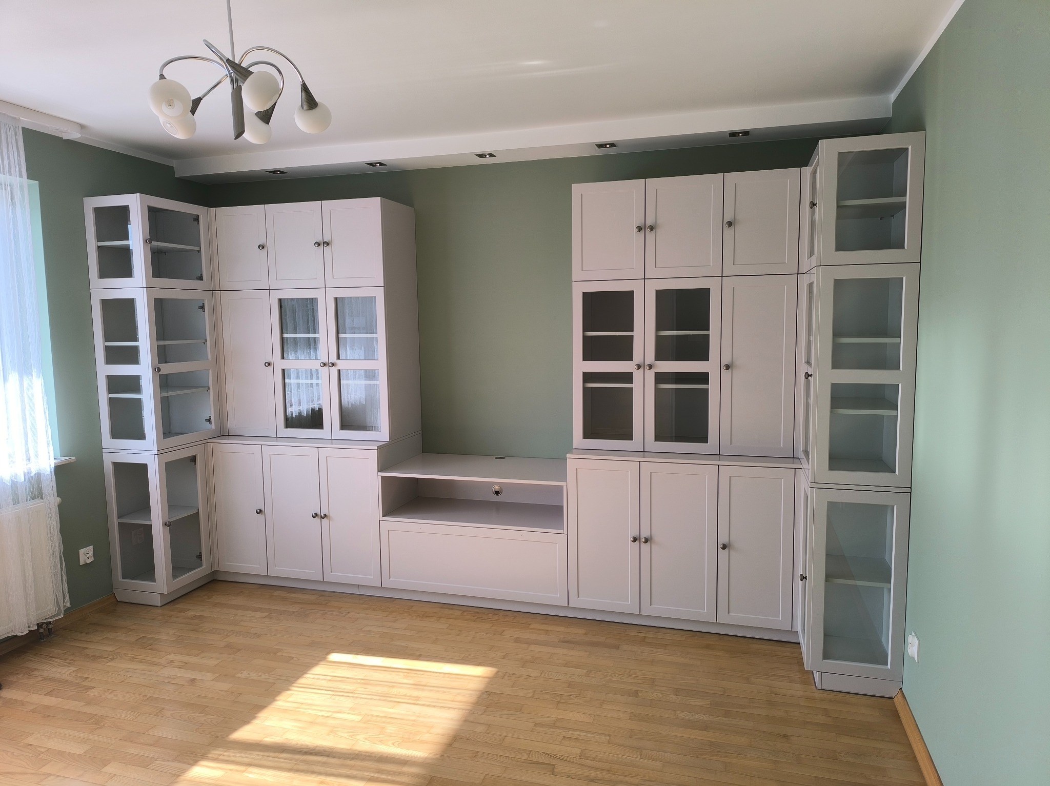 Jasna zabudowa meblowa z przeszklonymi witrynami i szafkami, dopasowana do wnętrza z drewnianą podłogą i zielonymi ścianami. Oświetlenie sufitowe.