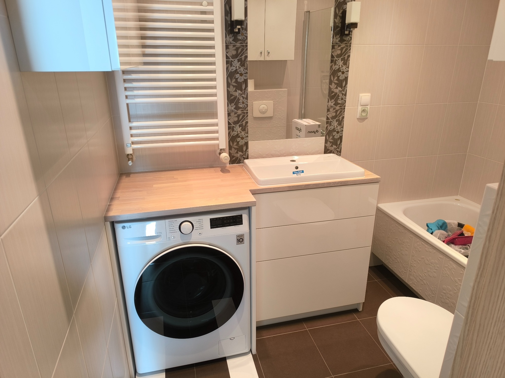 Nowoczesna łazienka z pralką LG w zabudowie, blat z jasnego drewna, biała umywalka z szafką i grzejnik drabinkowy. Beżowe płytki, minimalistyczny design, funkcjonalne rozwiązanie.